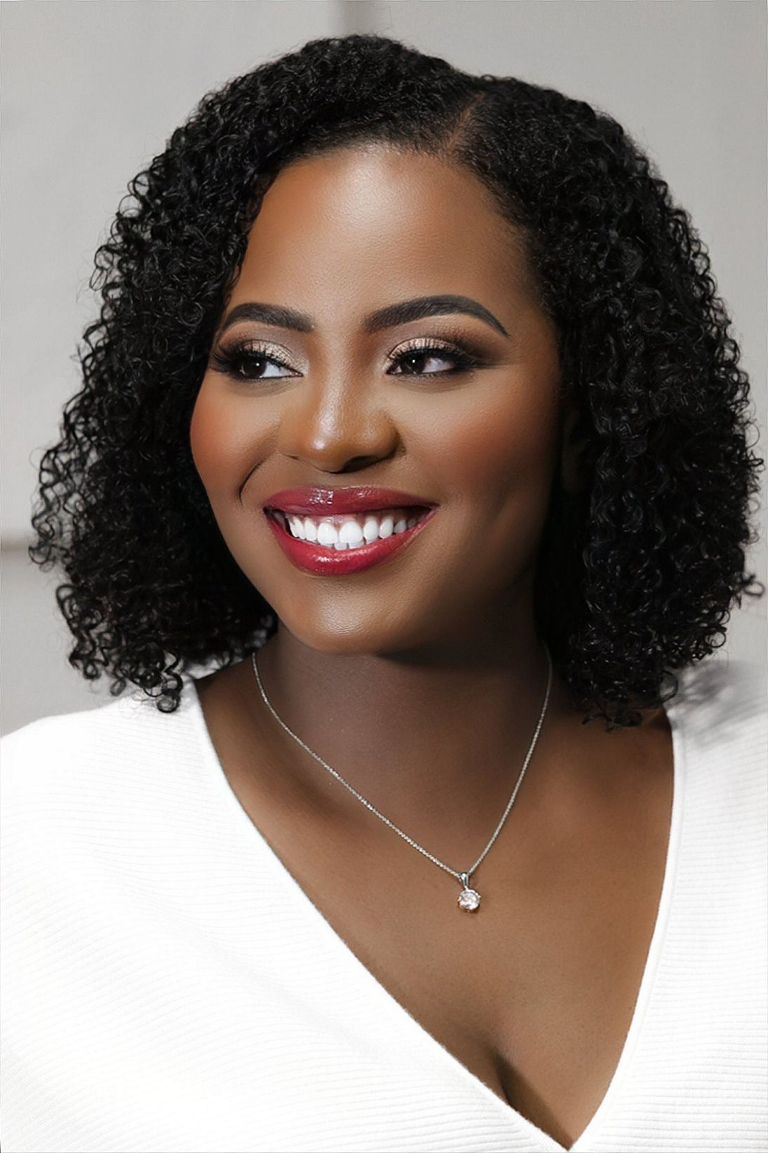 Confident Black woman with curly hair wearing white top and silver necklace, bright smile.