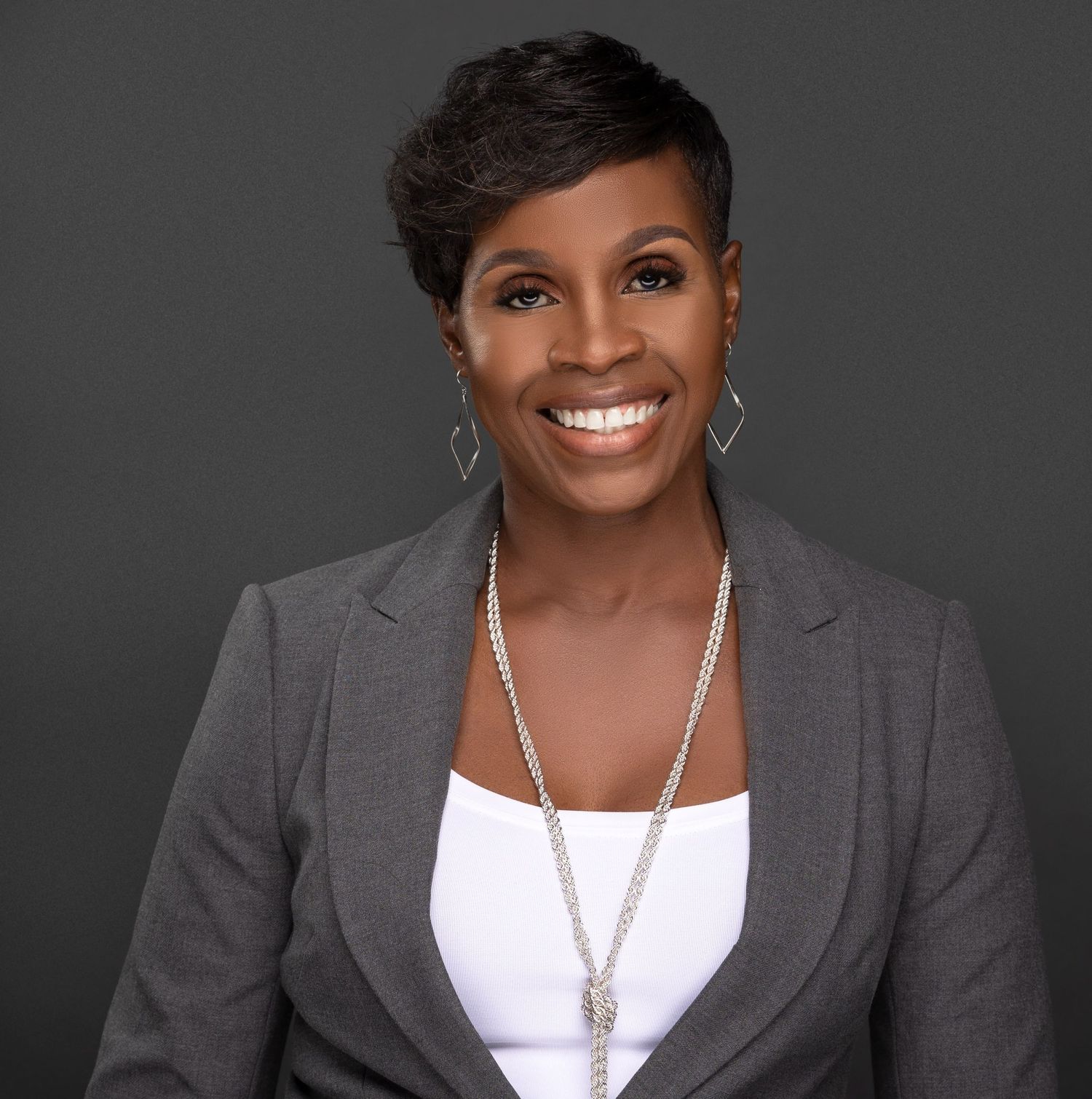 Professional woman in gray blazer smiling confidently against dark background.