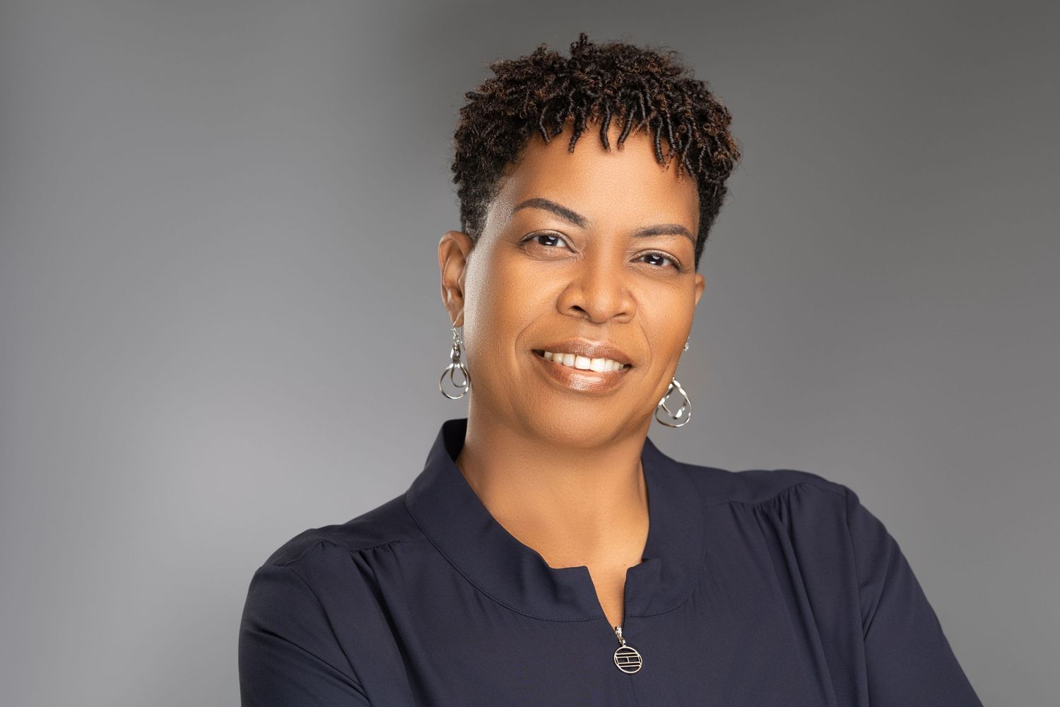 Smiling Black woman with short natural hair in navy top poses for professional headshot on gray backdrop.