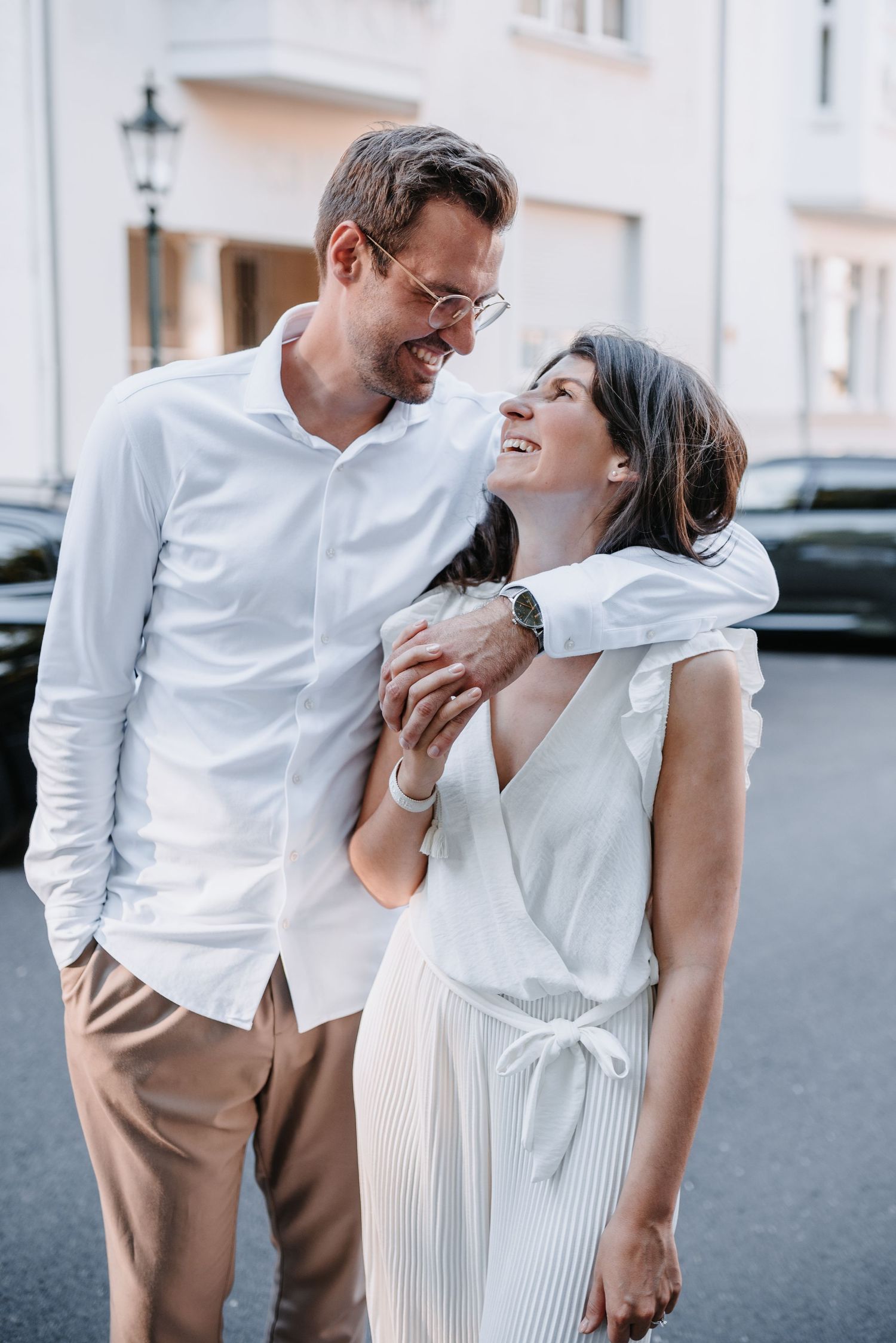A couple in matching white outfits share a candid laughing moment on a city street.