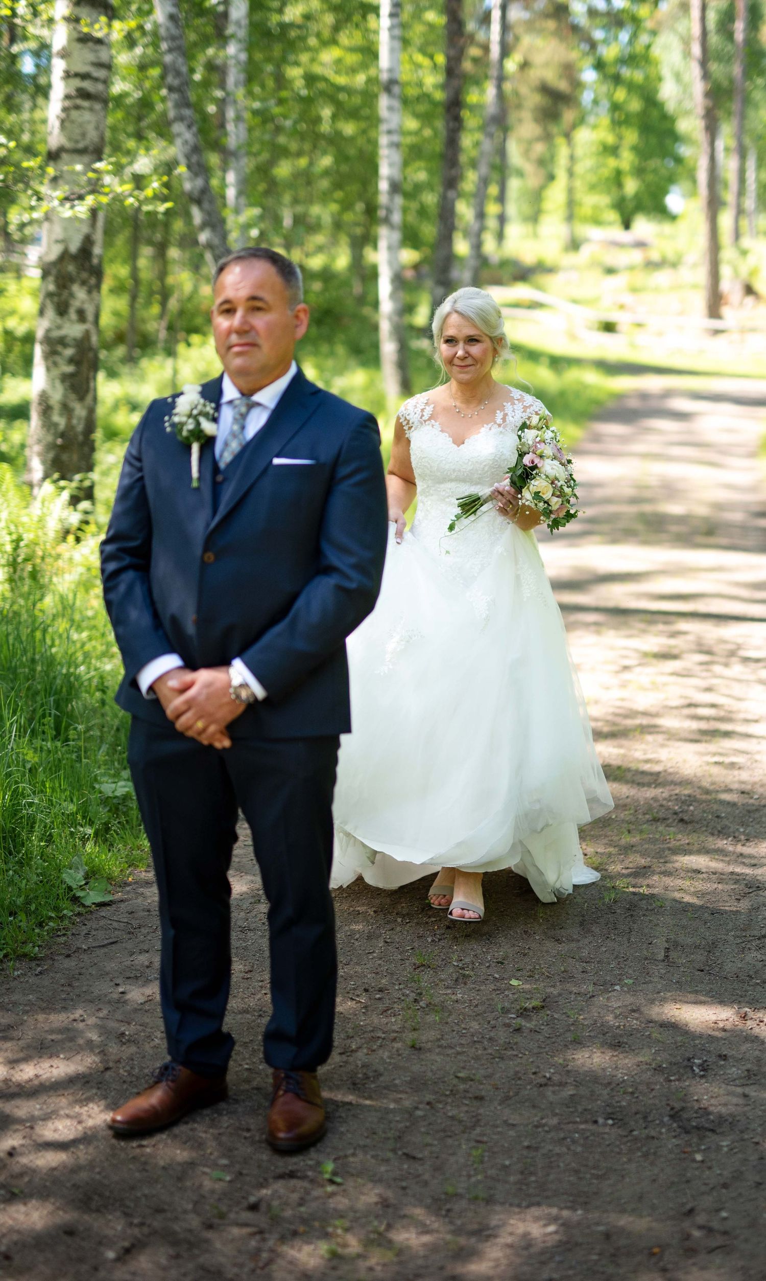 Groom stands on a forest path as his bride in a white lace gown approaches carrying a floral bouquet.