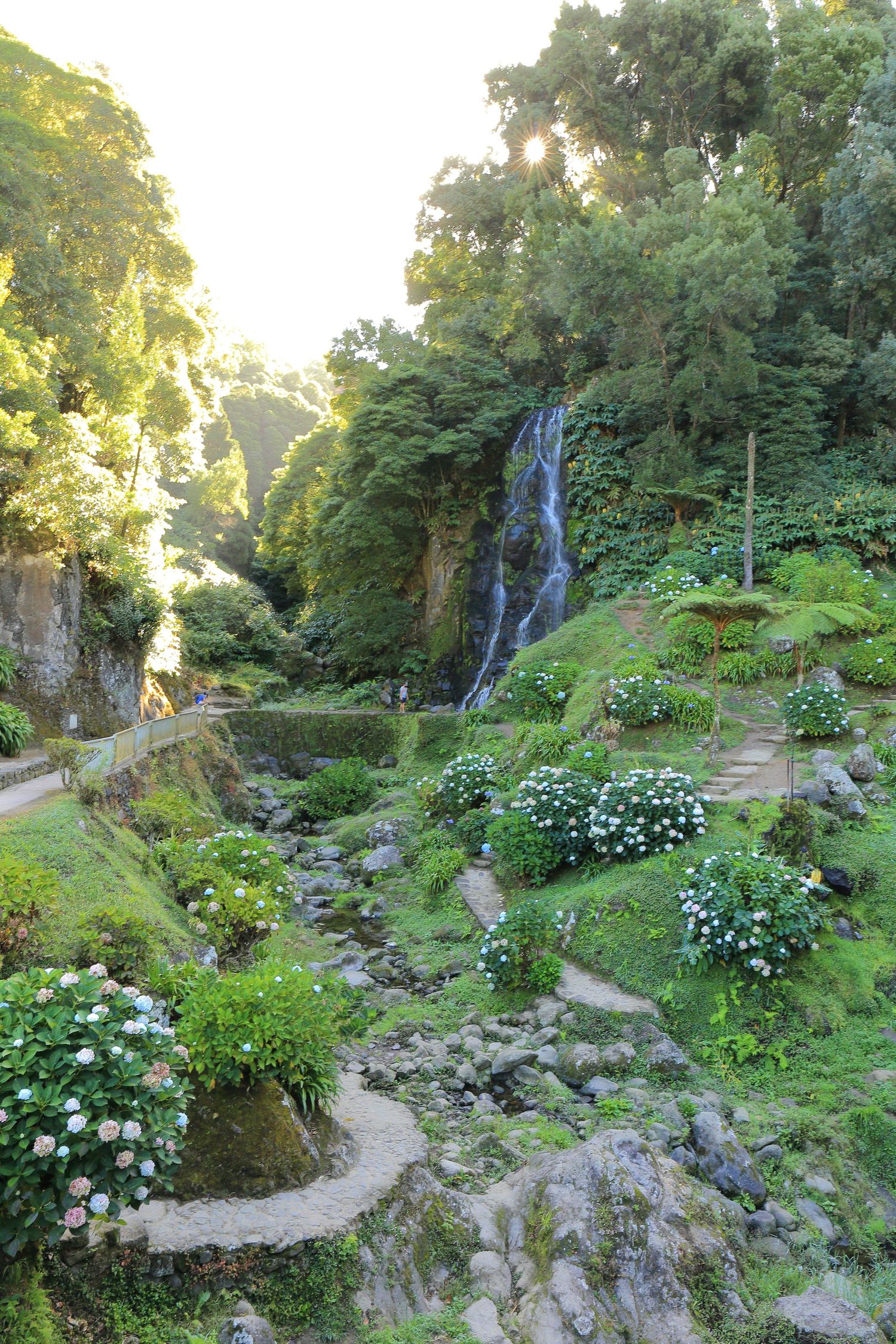 Scenic waterfall cascades down lush green cliffs in a manicured garden setting with winding paths.