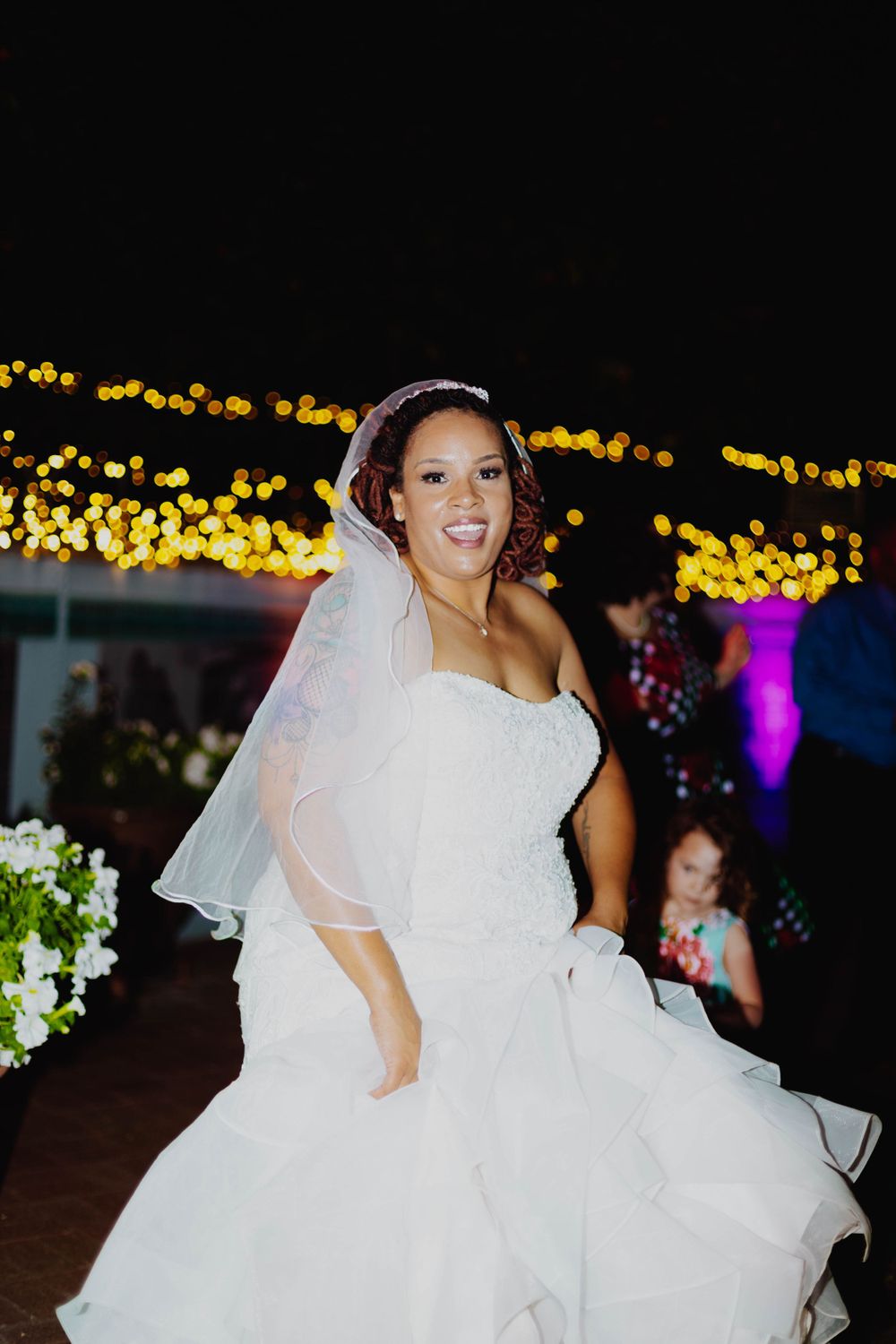 Bride in strapless white gown twirls on dance floor with decorative lights in background.