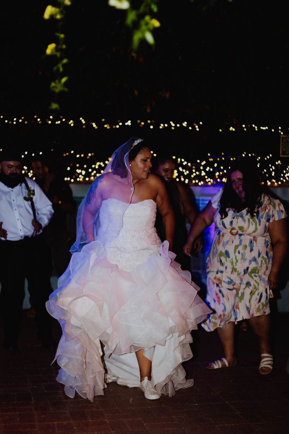 Bride in flowing white dress dances joyfully during wedding reception under twinkling lights.