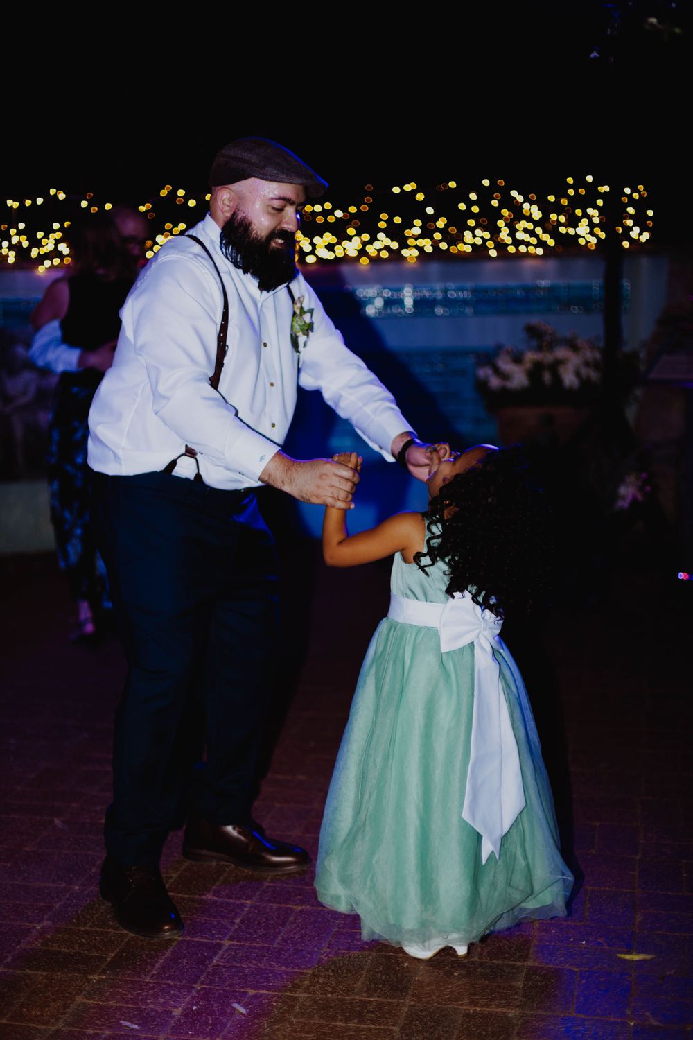 Man in suspenders dances with young girl in mint green dress at wedding reception.