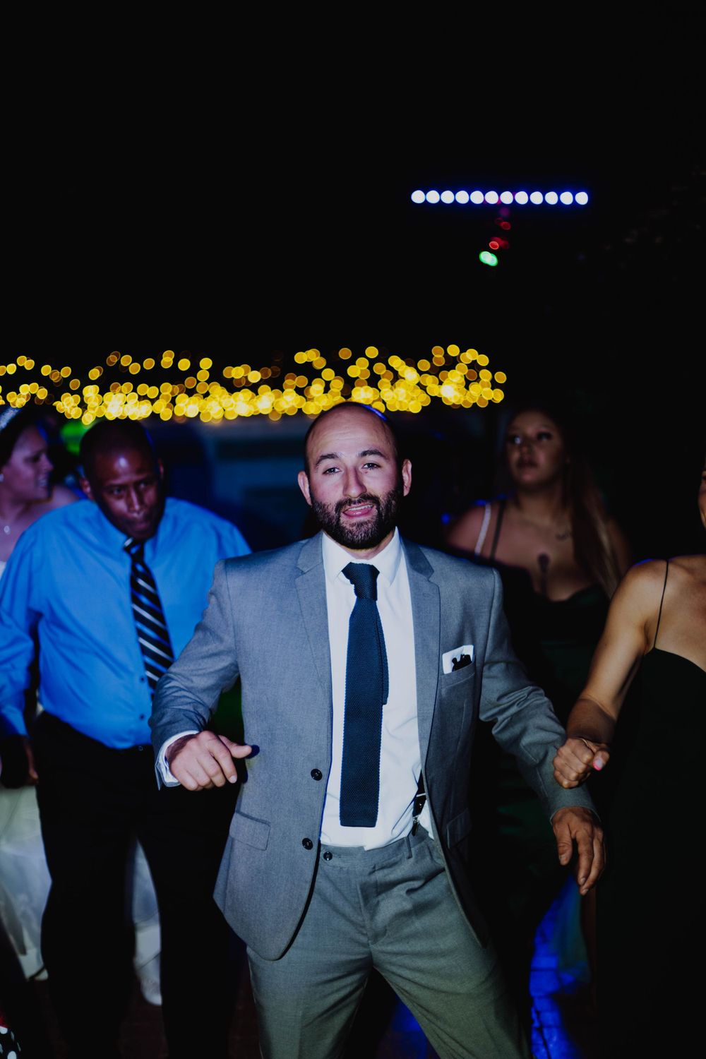 Wedding guest in grey suit dances enthusiastically at reception with bokeh lights in background.