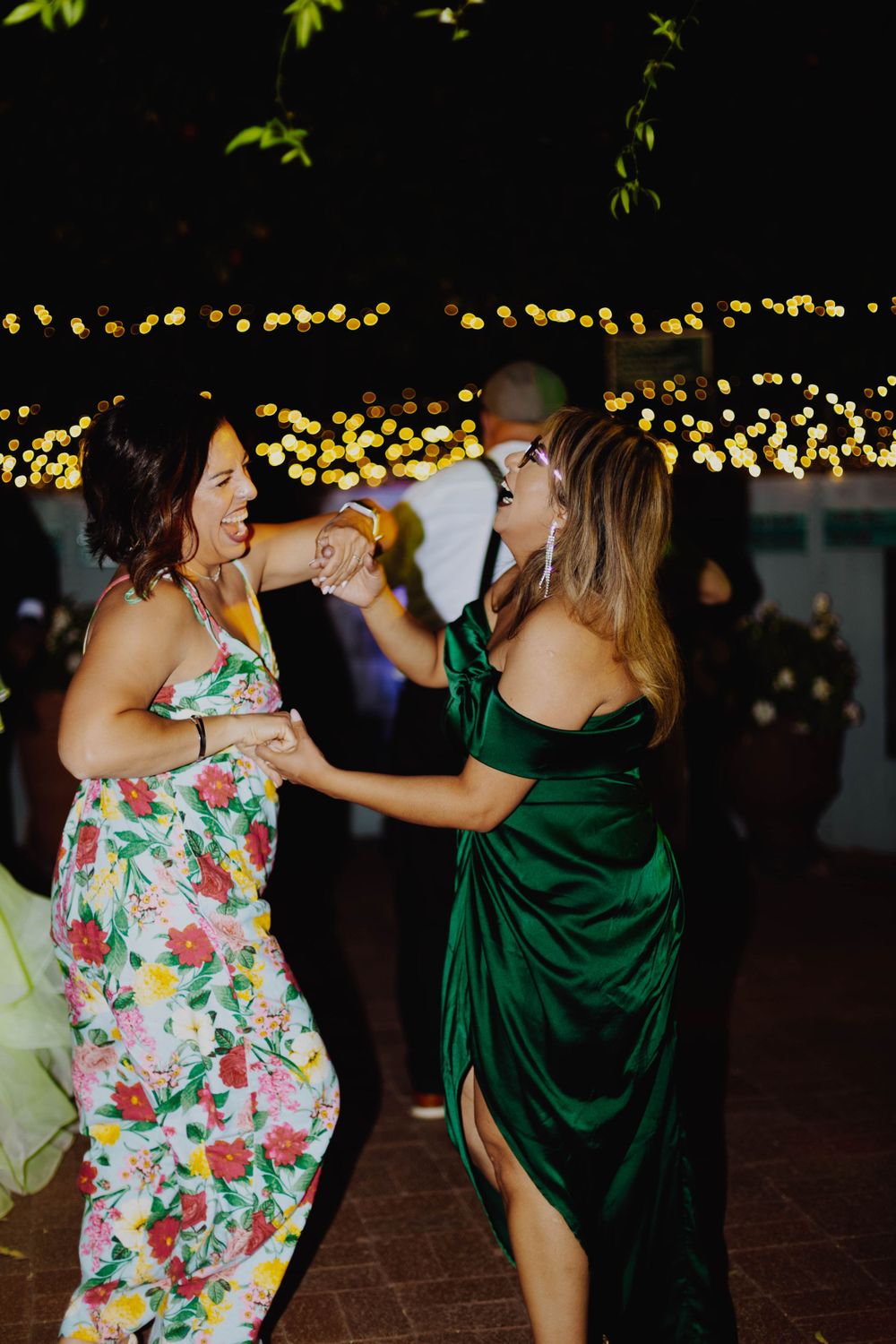 Two wedding guests dance together, one in floral dress and other in emerald green gown.
