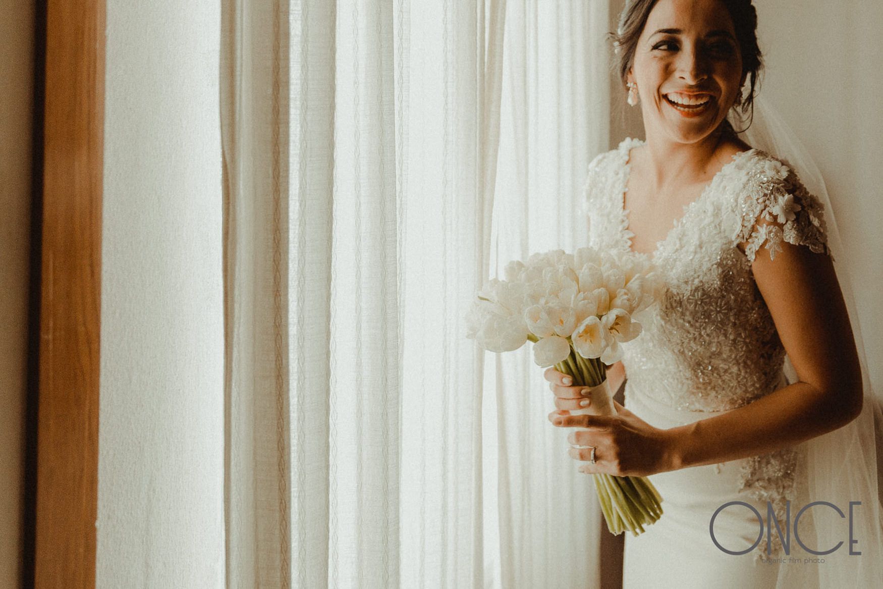 A delicate white bridal bouquet held against soft window light and sheer curtains.