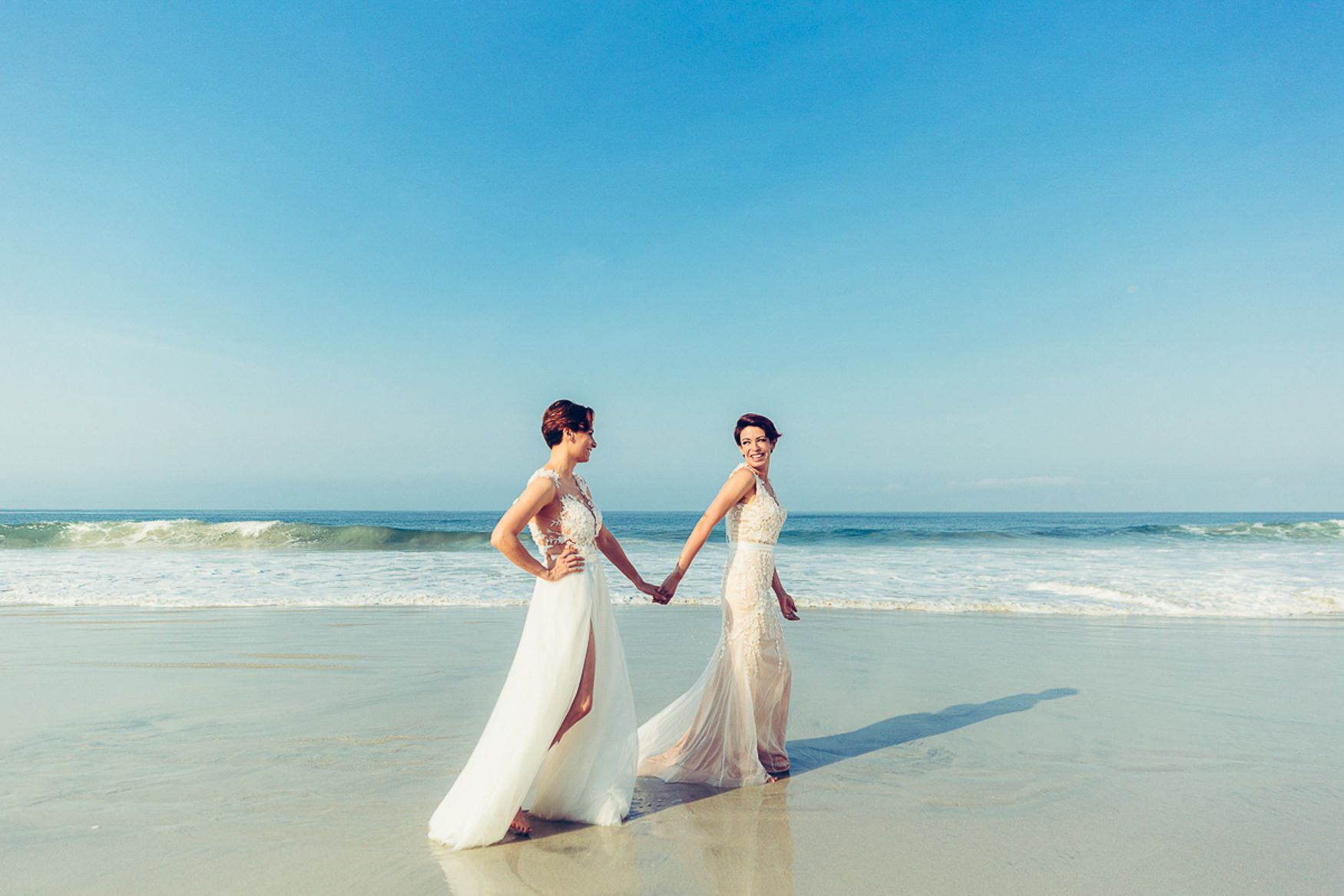 Two white dresses flowing in the ocean breeze on a pristine beach with clear blue skies.