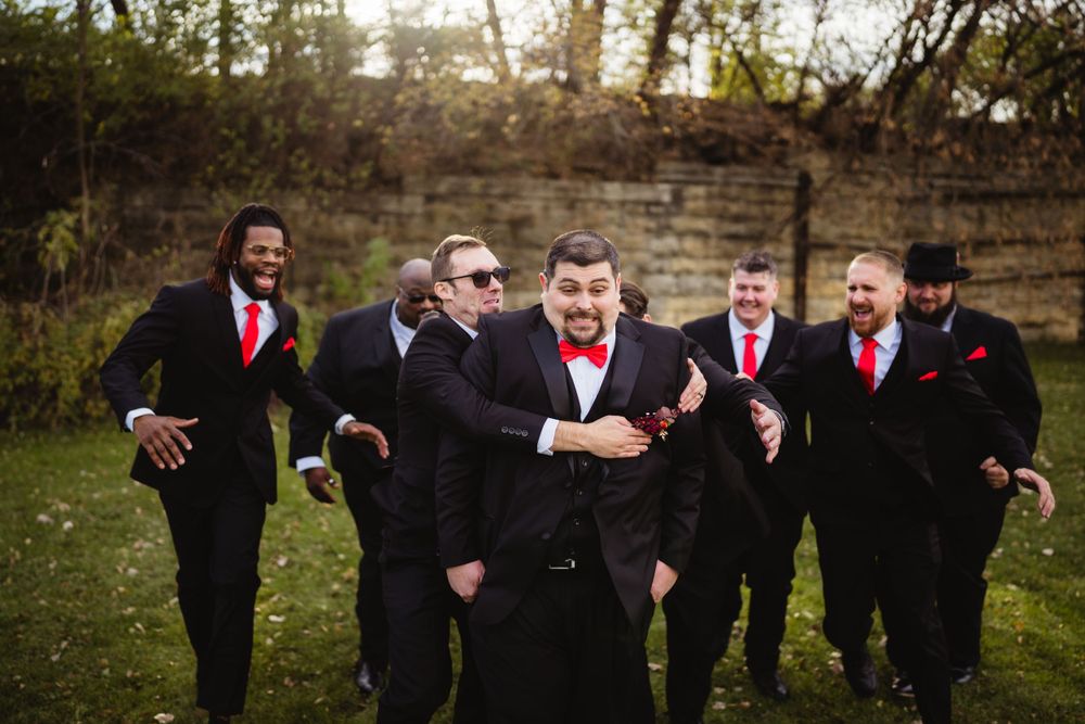 Groomsmen in black suits with red accents pose together outdoors in a playful moment.