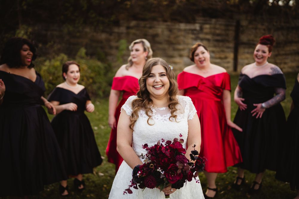 Bridal party in alternating black and red dresses pose by stone wall.