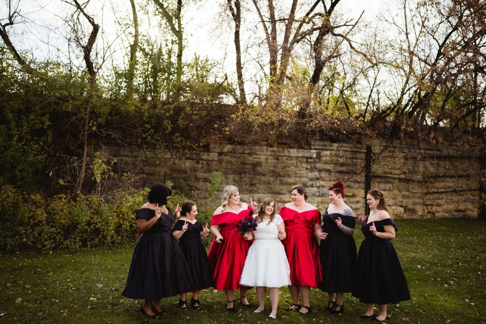 Wedding party in red and black dresses lined up against stone wall outdoors.