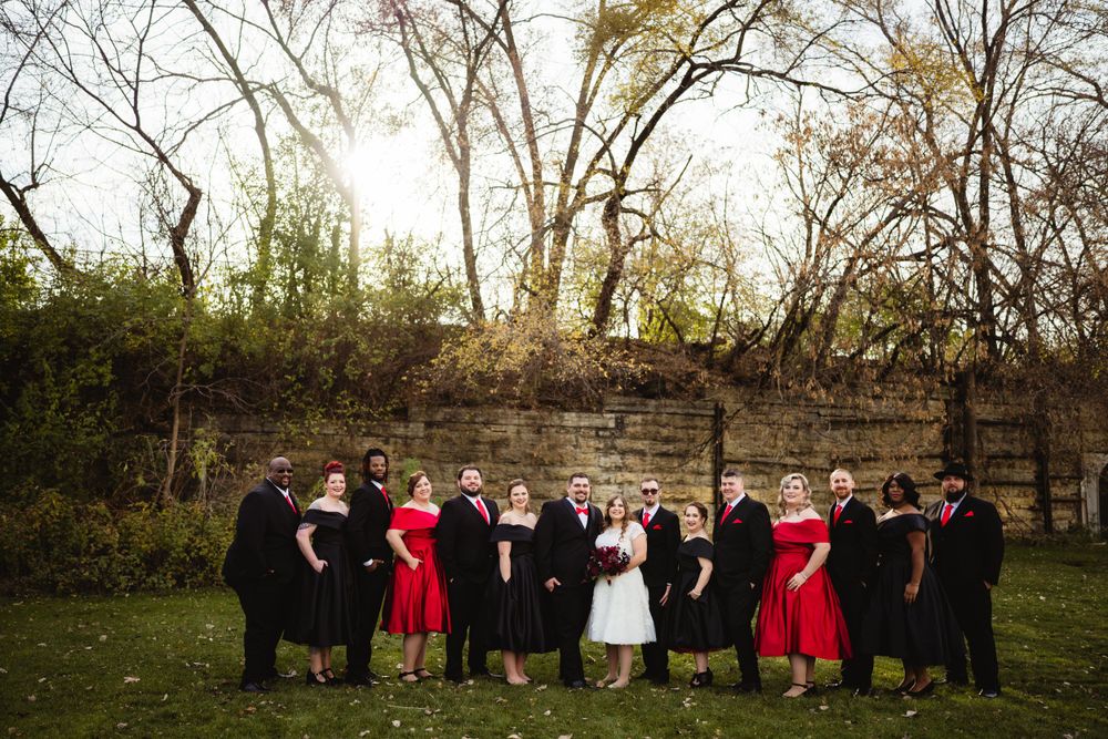 Wedding party in black and red attire poses together against stone wall at sunset.