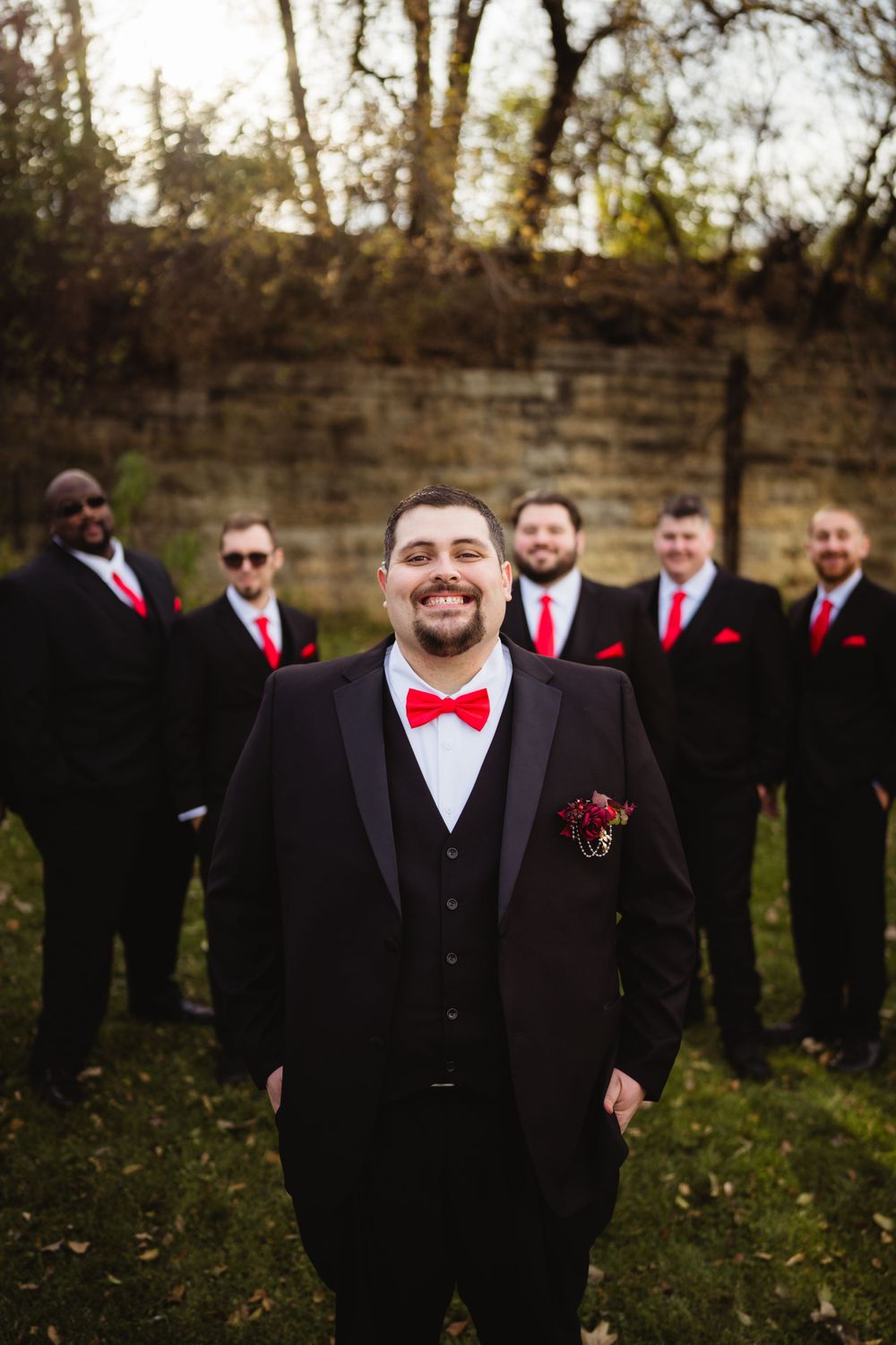 Groom with red bowtie poses with groomsmen in black suits against stone wall.
