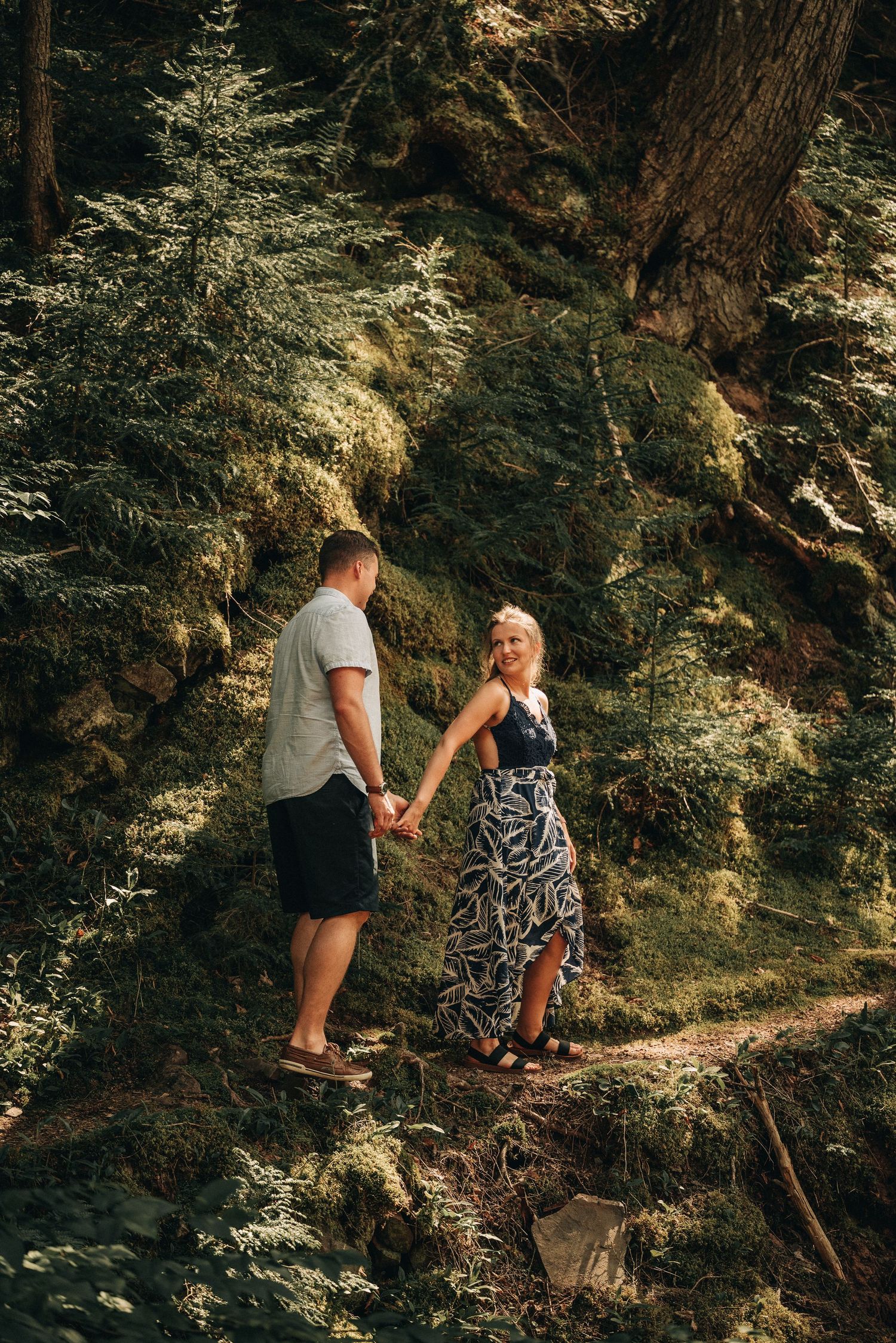 A couple stands on a forest trail with sunlit moss-covered rocks in the background.