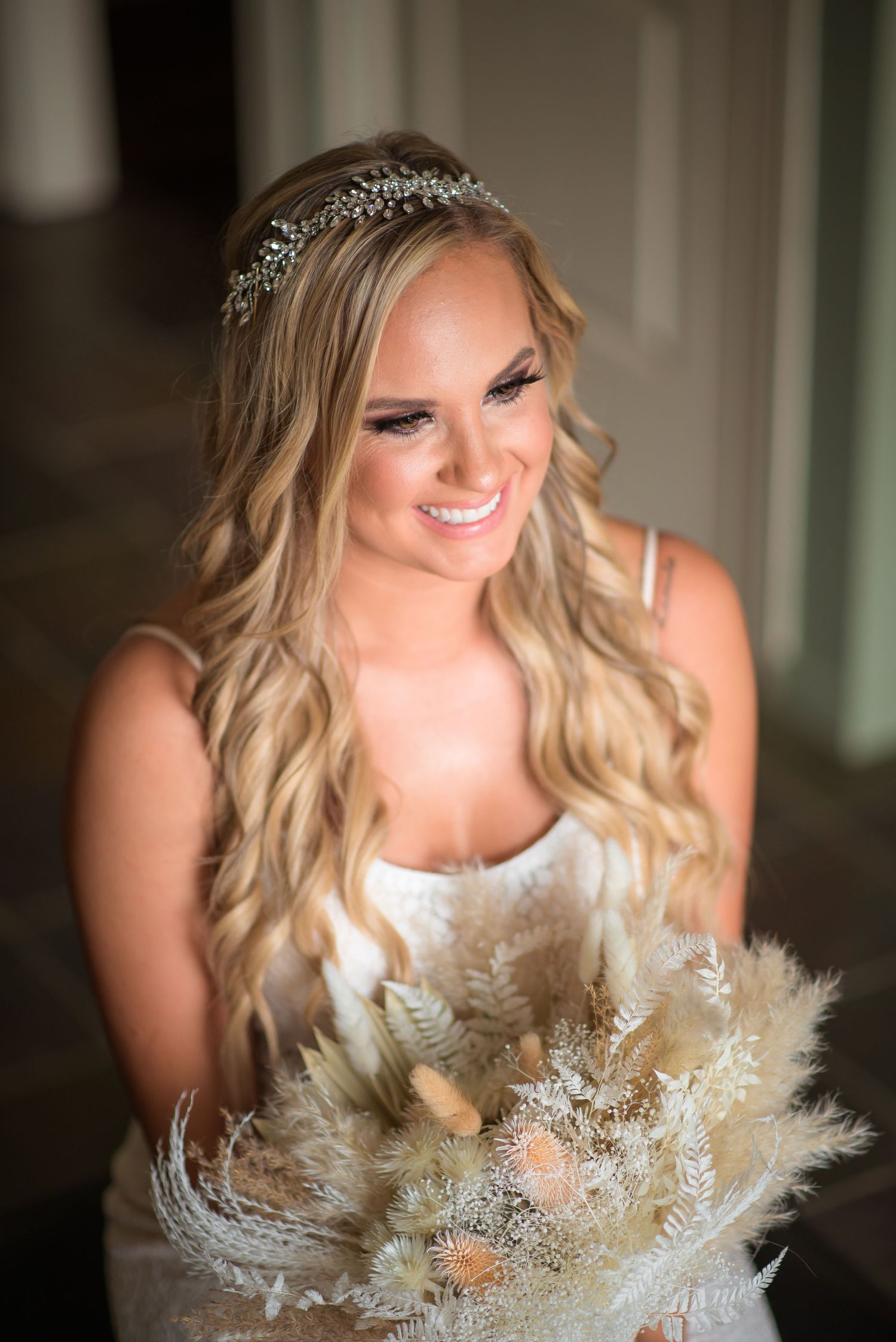 Smiling blonde bride wearing a crystal crown holding a boho dried floral bouquet on her wedding day.