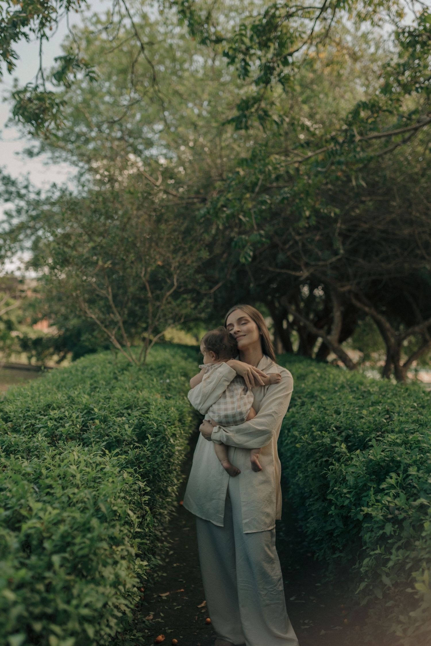 Parent embracing baby while standing in a lush garden path surrounded by greenery.