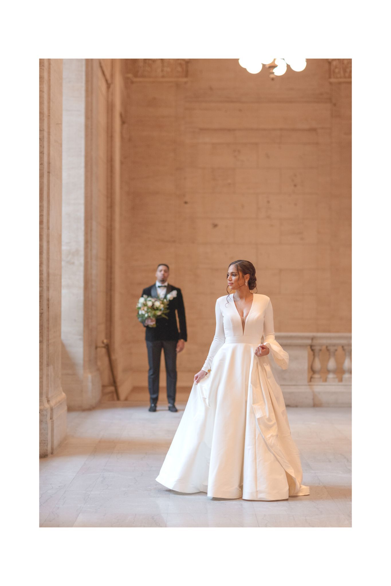Couple in wedding attire pose elegantly in historic building with stone columns.