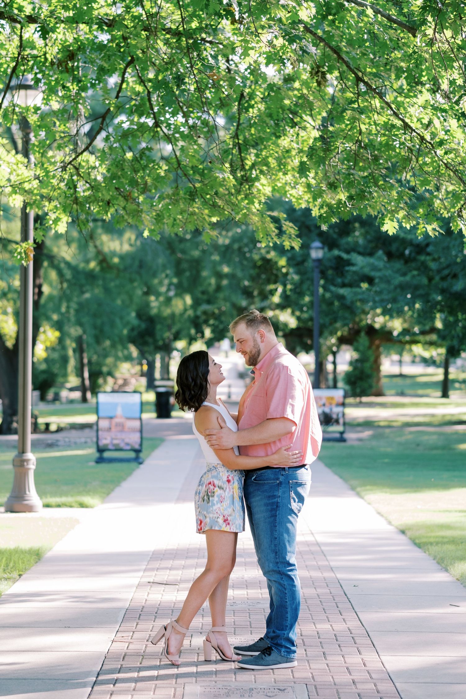 Romantic engagement shoot on a tree-lined pathway in a summer park.