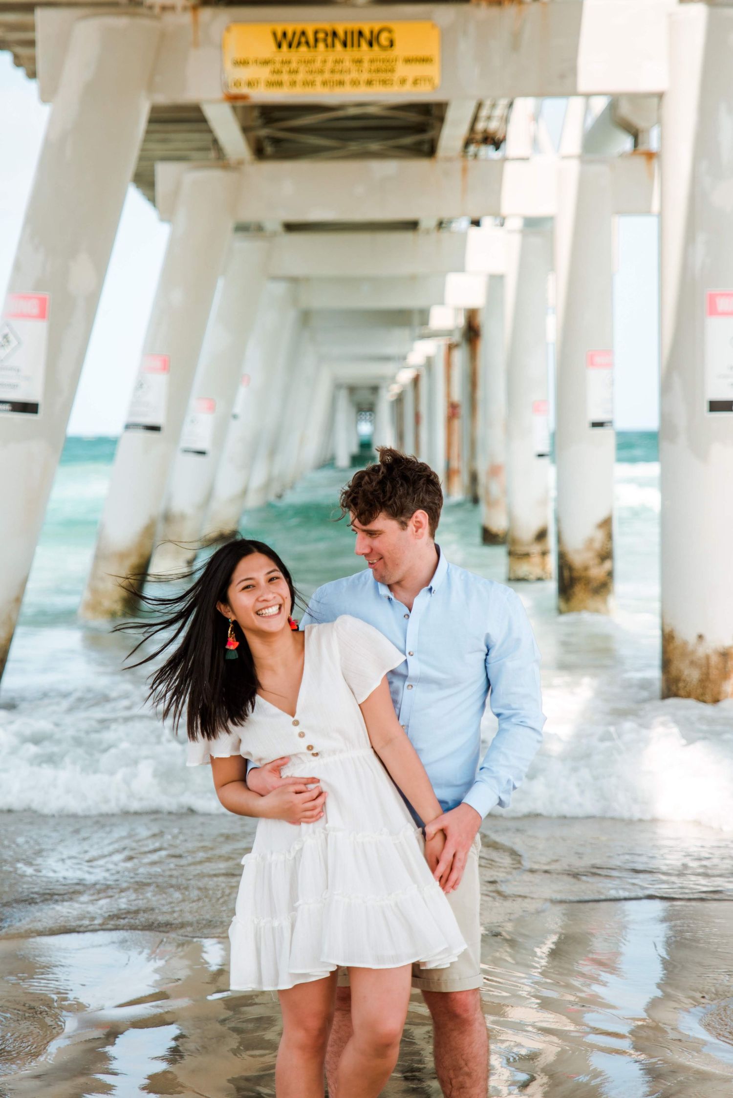 A joyful couple embracing under a pier with ocean waves swirling around their feet.