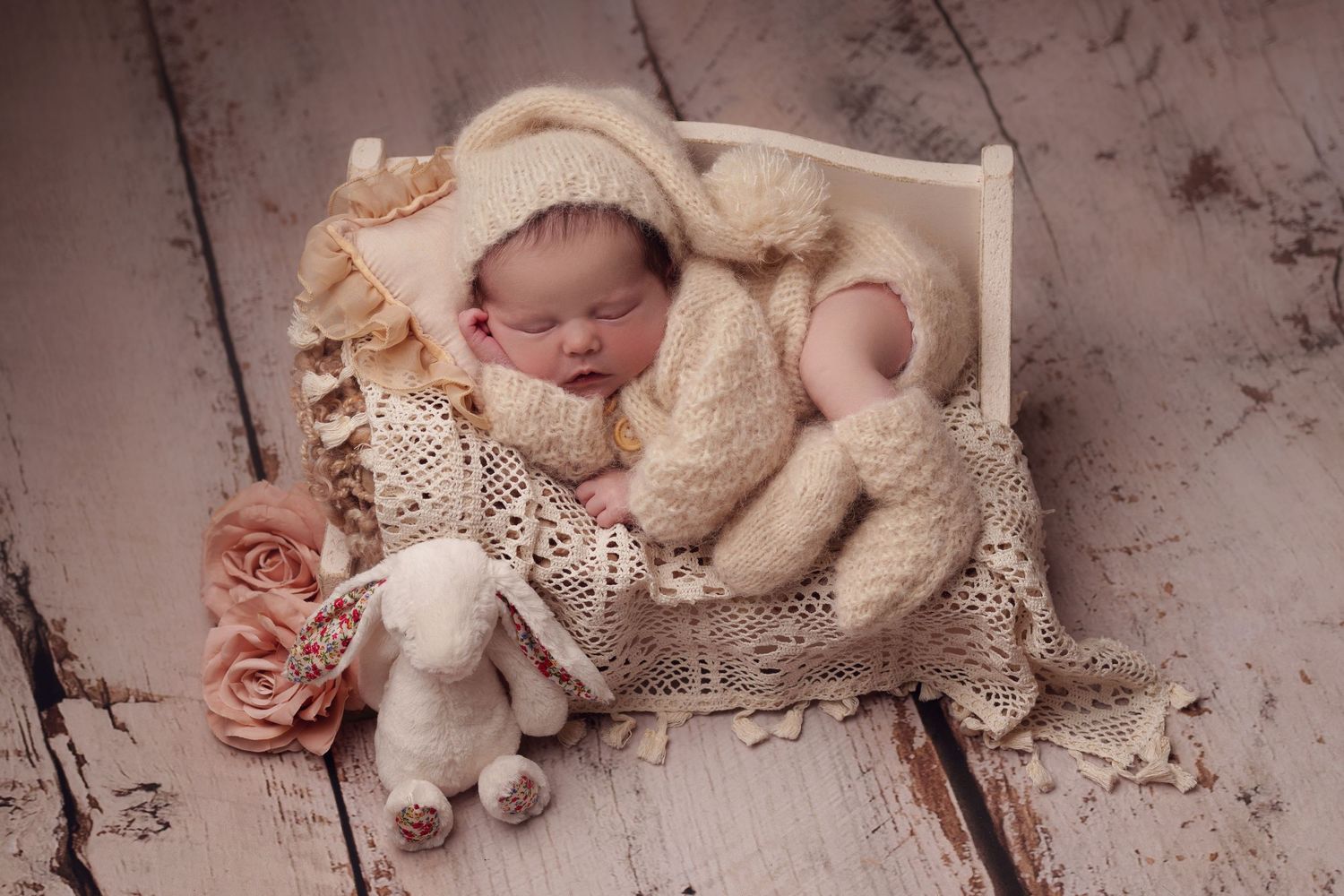 A peaceful sleeping newborn wearing a cream knit outfit rests on white lace bedding with soft pink roses.
