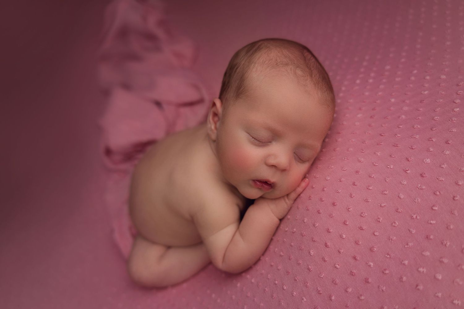 A sleeping newborn baby rests peacefully on a soft pink textured blanket in a professional portrait setting.