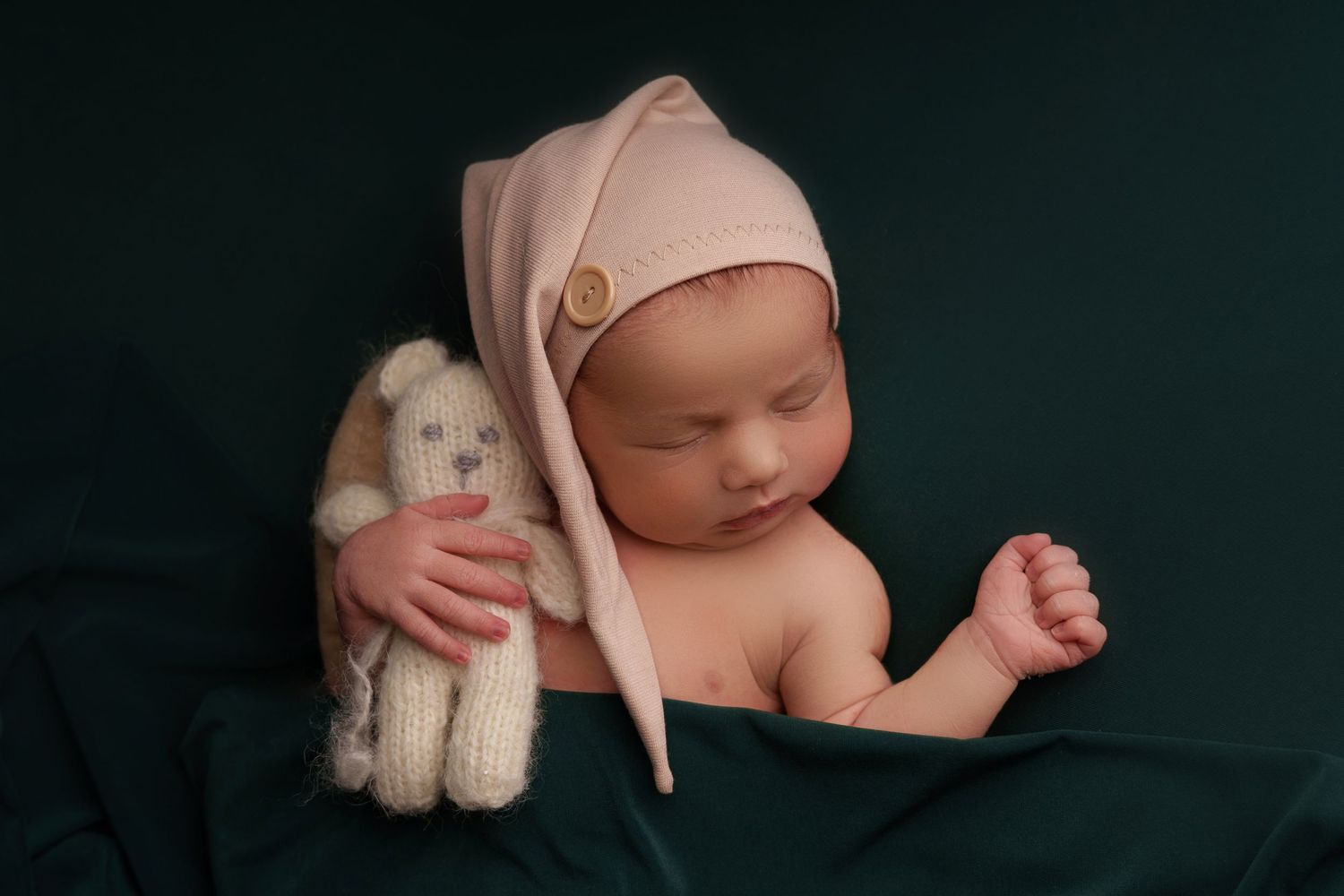 A sleeping baby in a pink bonnet lies on dark fabric with a plush white stuffed animal bunny.