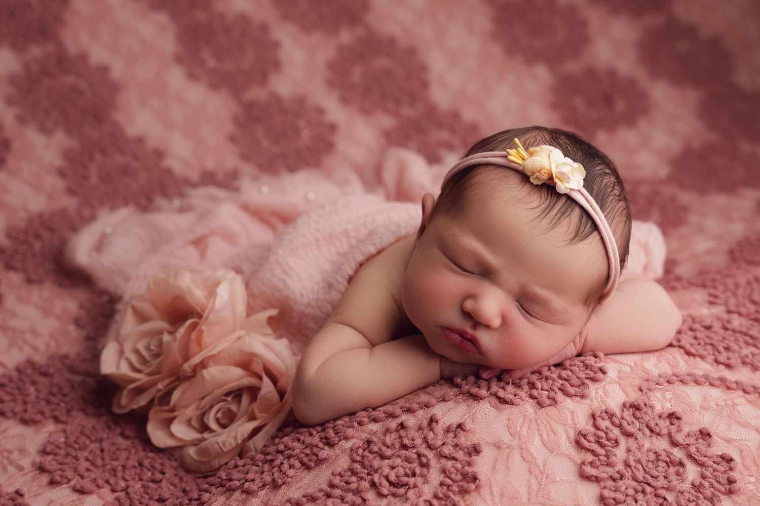 Sleeping newborn on pink textured blanket with matching floral headband and rose decorations.