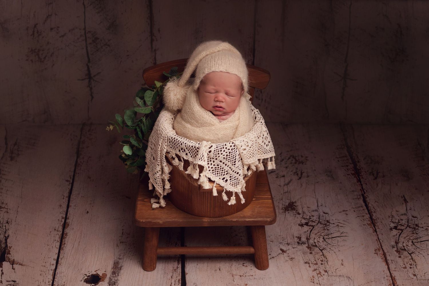 Sleeping newborn wrapped in cream knit blanket rests on wooden stool with greenery backdrop during professional photoshoot.