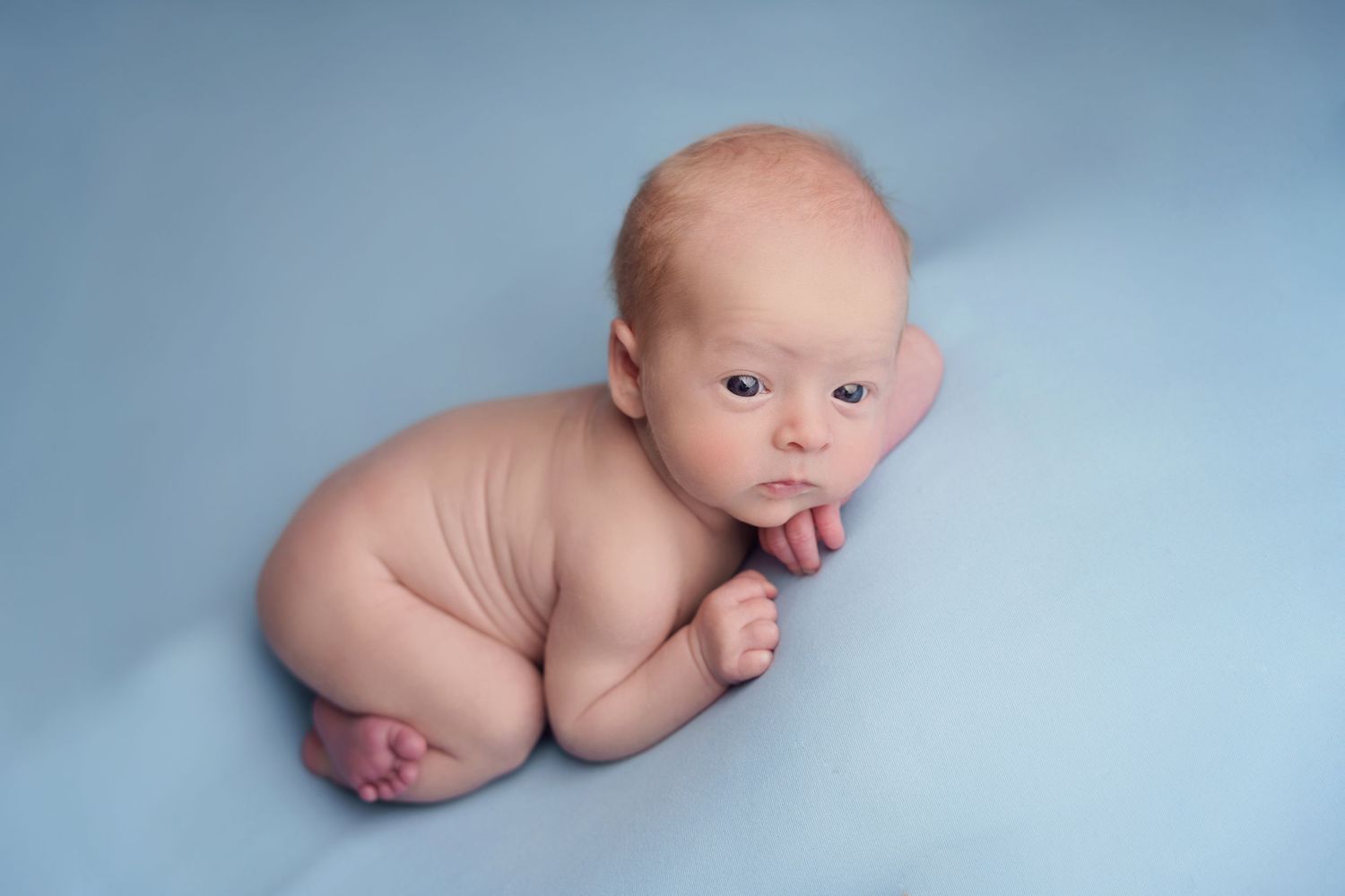 A newborn baby lying on a soft blue backdrop with their hand near their mouth.