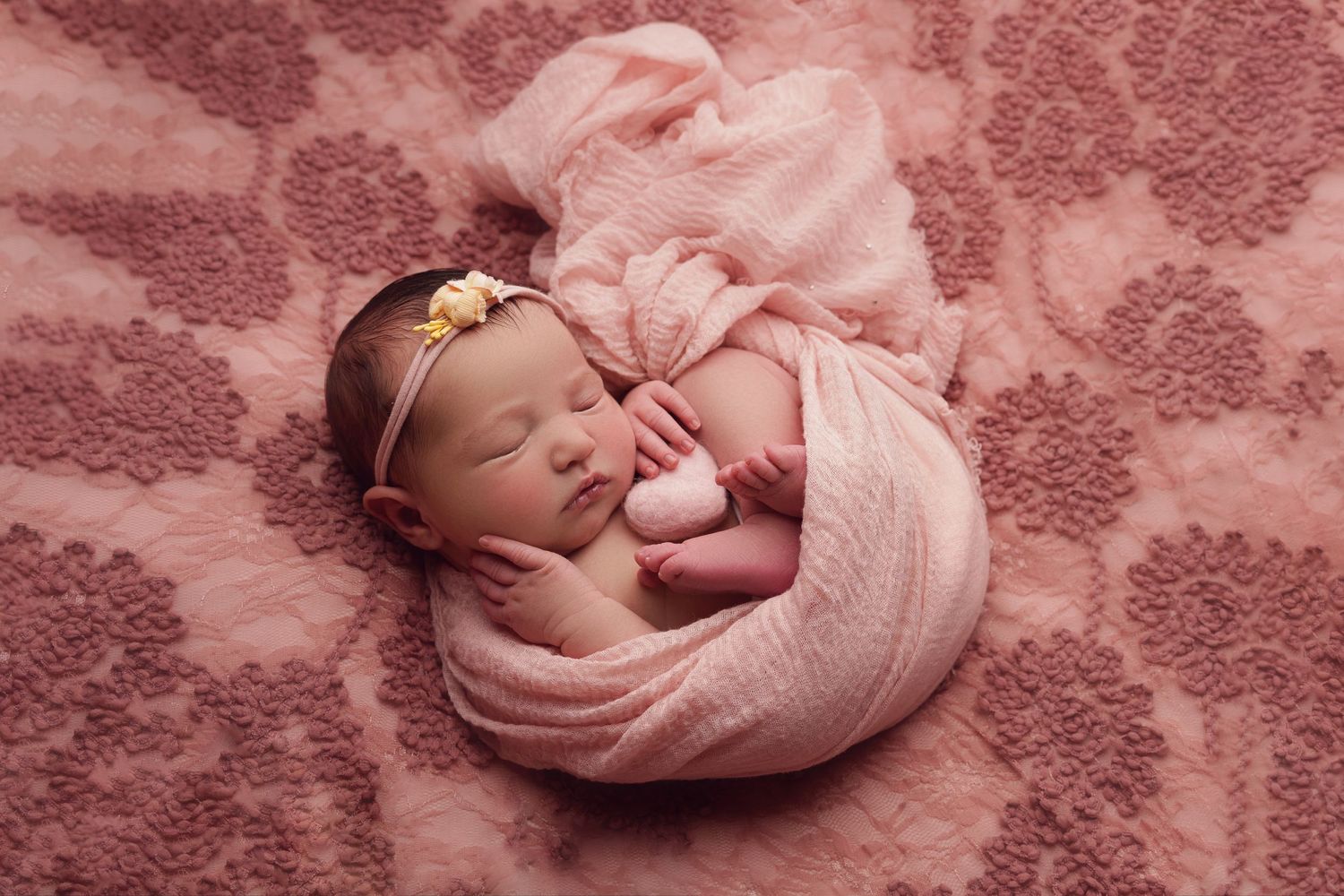 Sleeping newborn baby swaddled in soft pink wrap on pink textured background with delicate headband accessory.