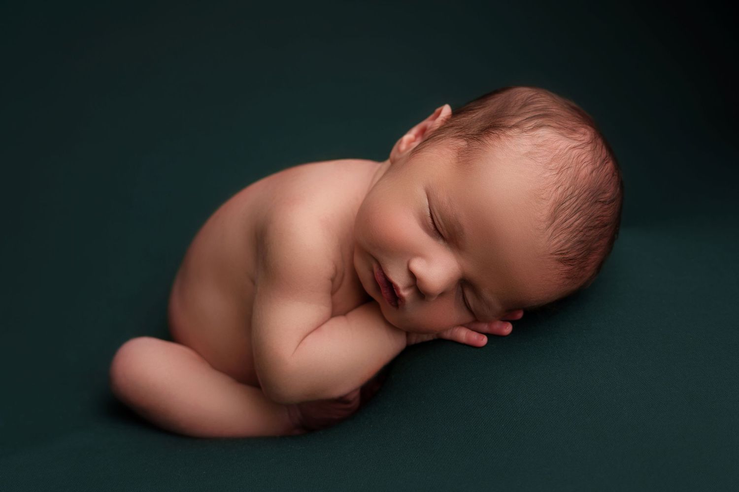 A peacefully sleeping newborn baby curled up on a dark green background in a minimalist portrait.