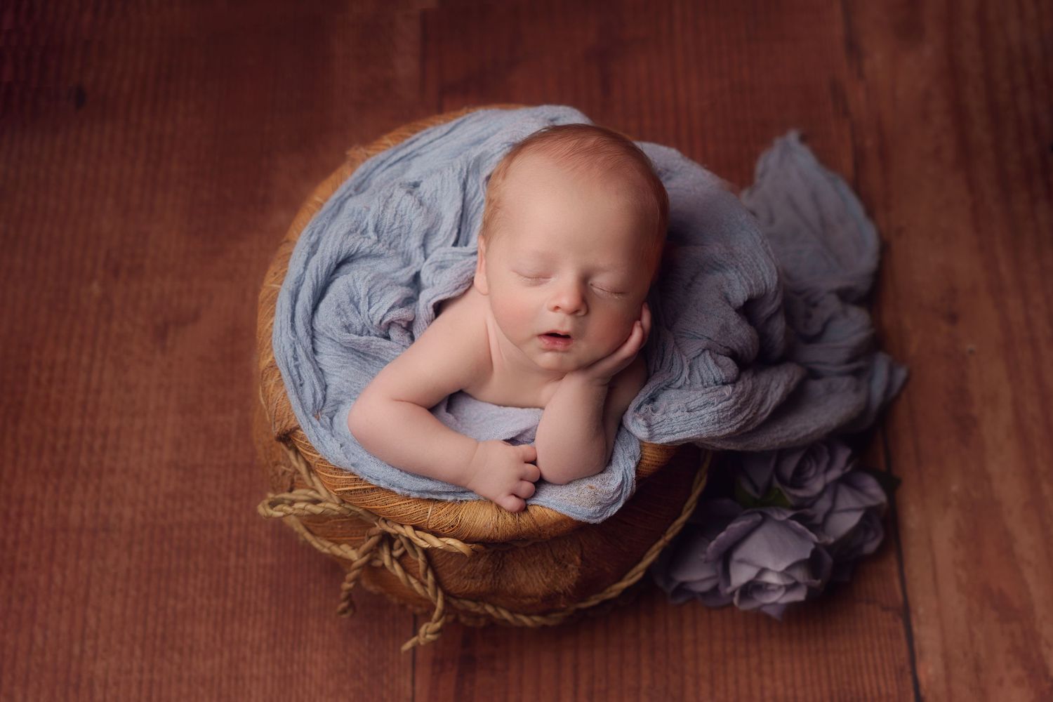 Sleeping newborn in blue fabric swaddling lies peacefully in rustic wooden bowl on brown surface.