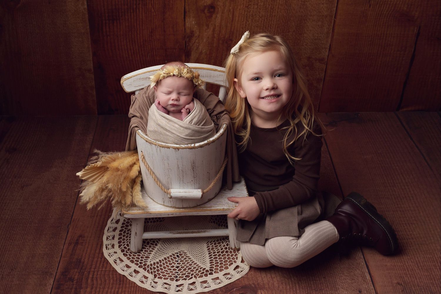 A baby nestled in a wooden bucket while a young child sits beside them on a wooden floor with lace decor.