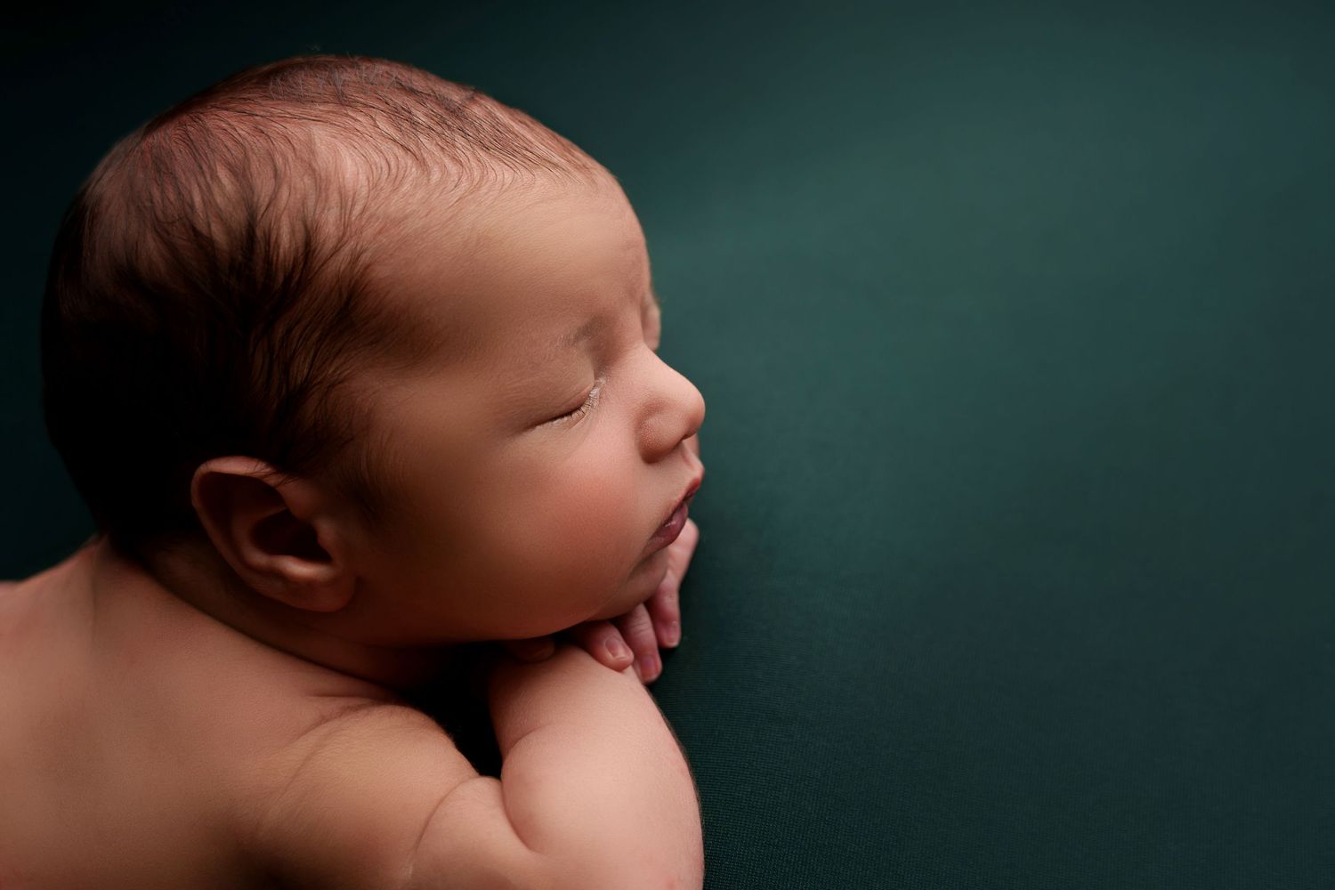 Peaceful sleeping newborn baby curled up against dark teal background in artistic portrait.
