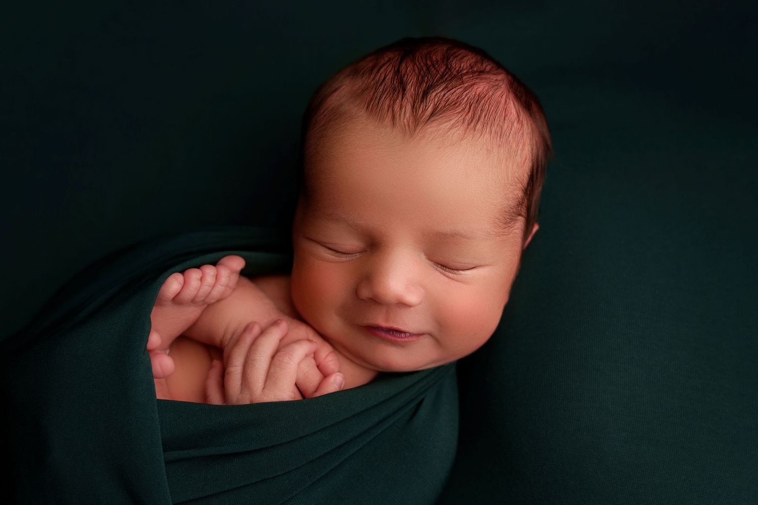 Sleeping newborn baby swaddled in dark green fabric against a black background, smiling peacefully.