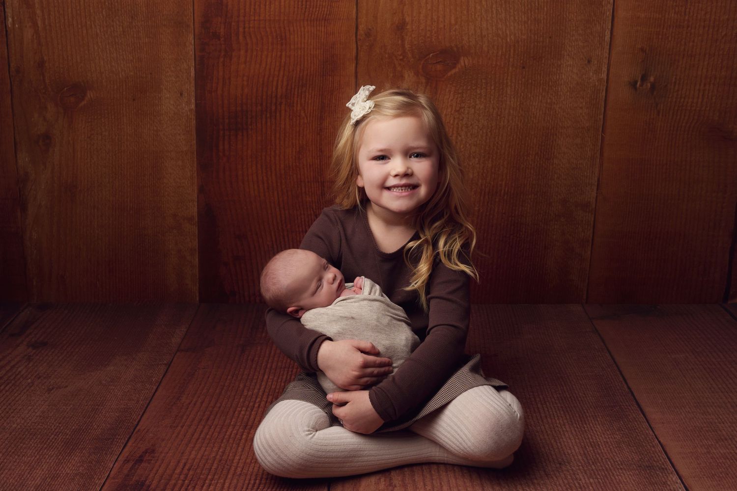 Young child in brown shirt sits cross-legged on wooden floor while holding a newborn baby.