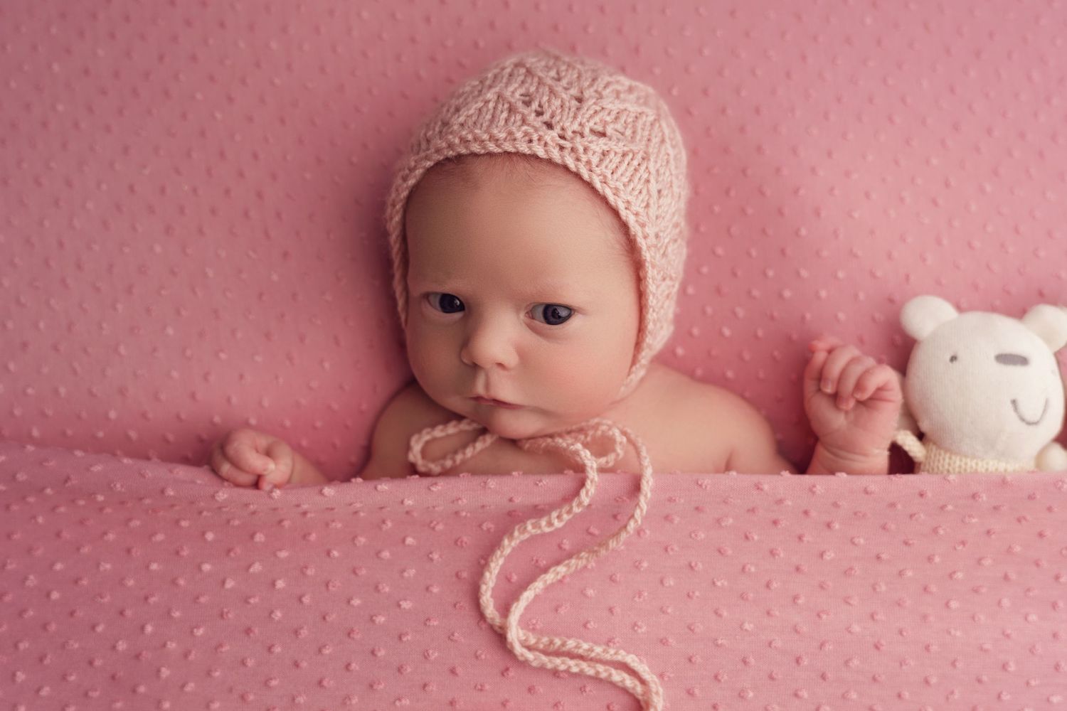 A baby wearing a pink knitted bonnet lies on pink textured fabric with a small white plush toy.