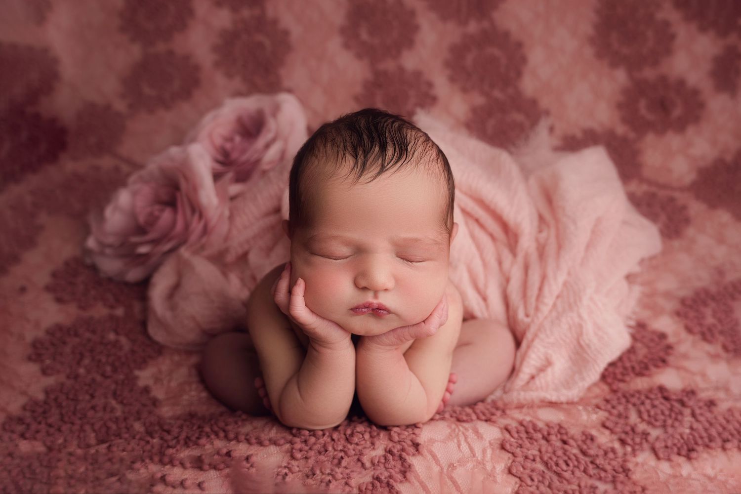 Peaceful newborn baby sleeping under pink blanket on soft textured rose-colored backdrop with floral pattern.