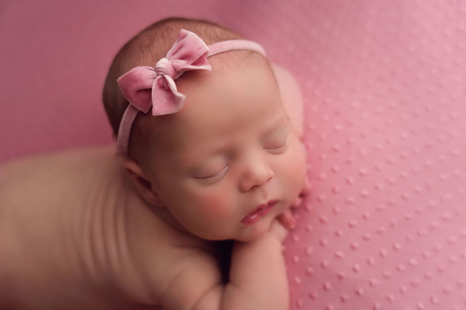 Peaceful sleeping newborn rests against pink dotted background wearing delicate bow headband.