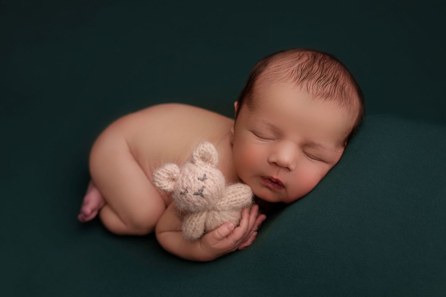 Peaceful sleeping newborn curled up with small teddy bear on dark green backdrop in professional portrait.