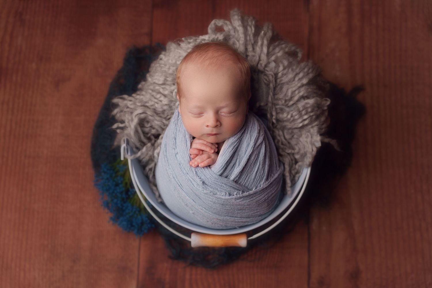 Newborn baby wrapped in soft gray fabric sleeping peacefully in a round navy and peacock blue nest on wooden floor.