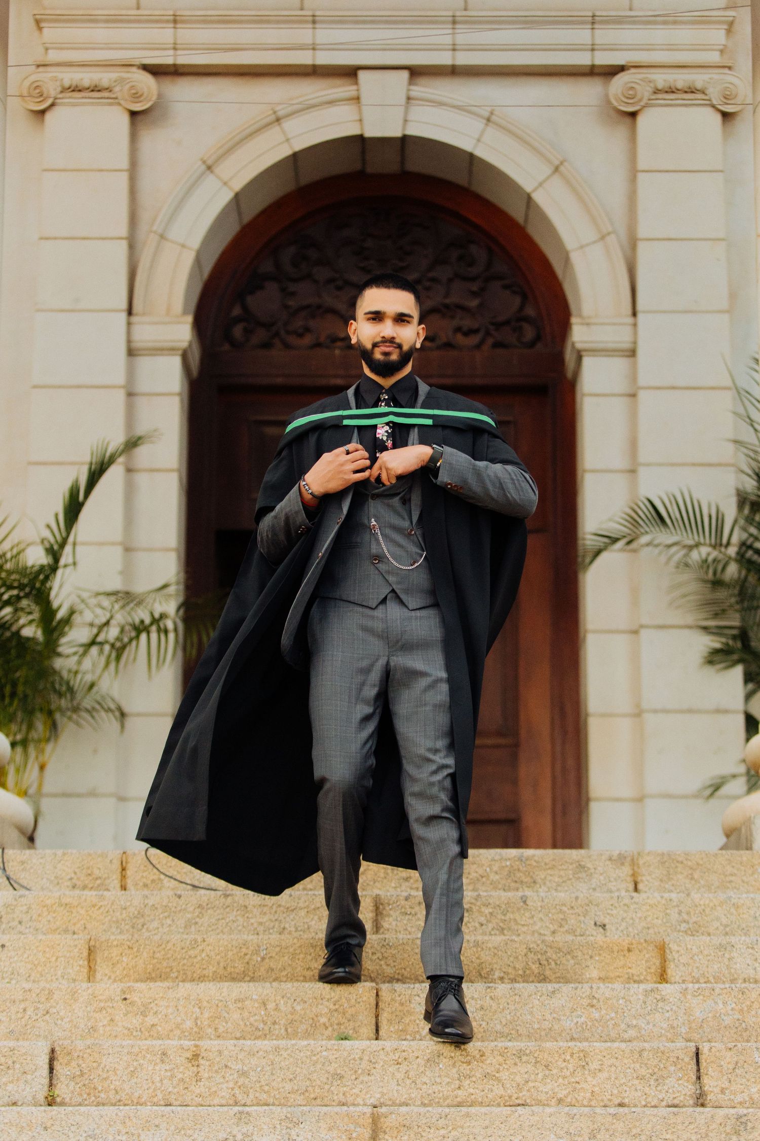 A graduate in a black academic gown and grey suit walks down stone steps in front of an ornate doorway with palm plants.
