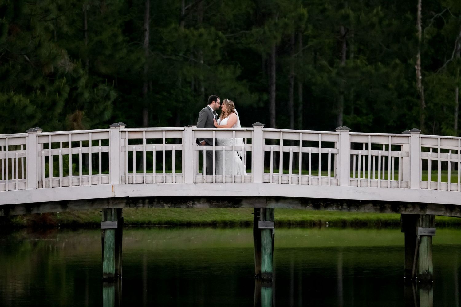 A couple shares a romantic moment on a white wooden bridge over a tranquil pond surrounded by green trees.