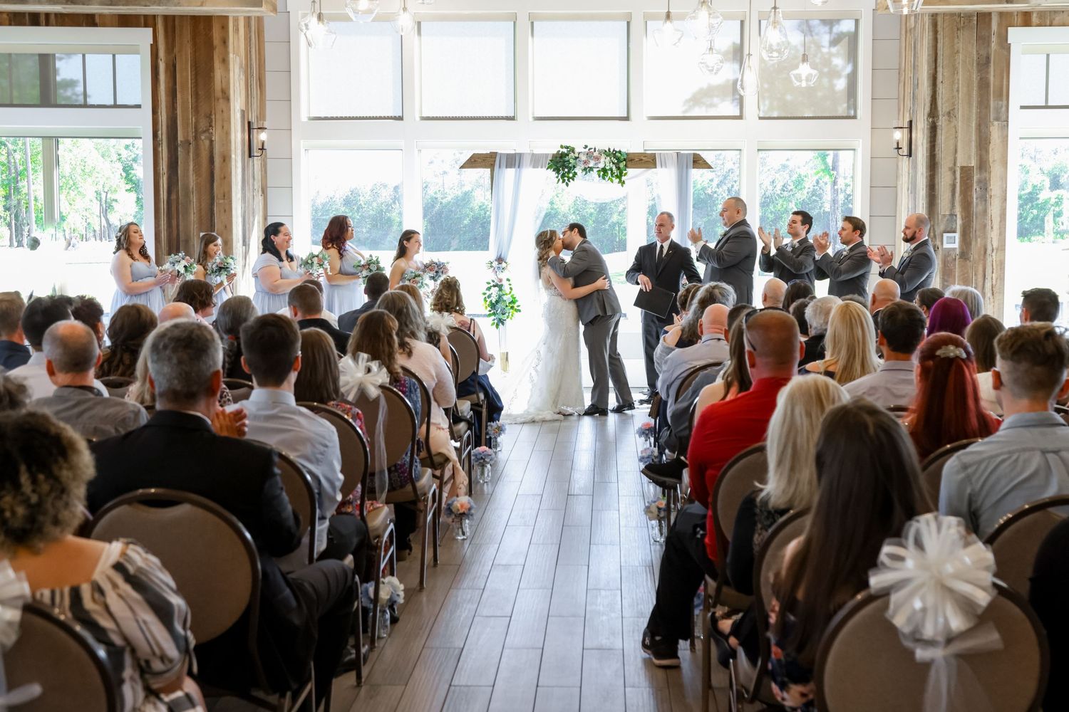 Indoor wedding ceremony with guests seated in wooden chairs watching couple exchange vows in natural light.