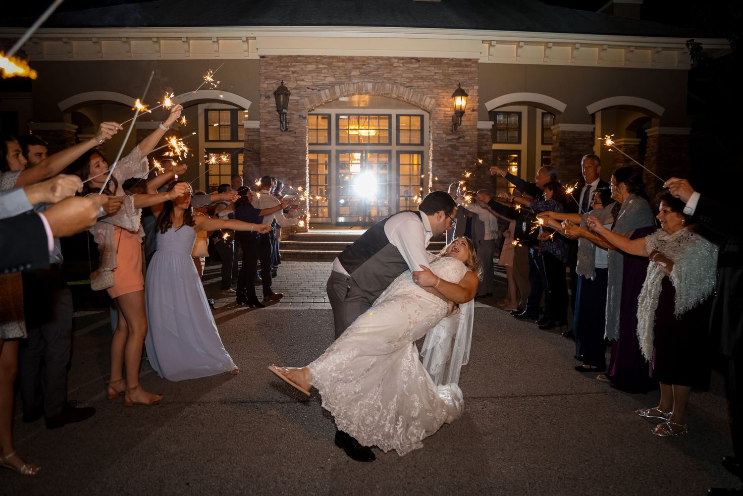 Wedding couple shares romantic dip kiss during sparkler exit at nighttime venue while guests celebrate.