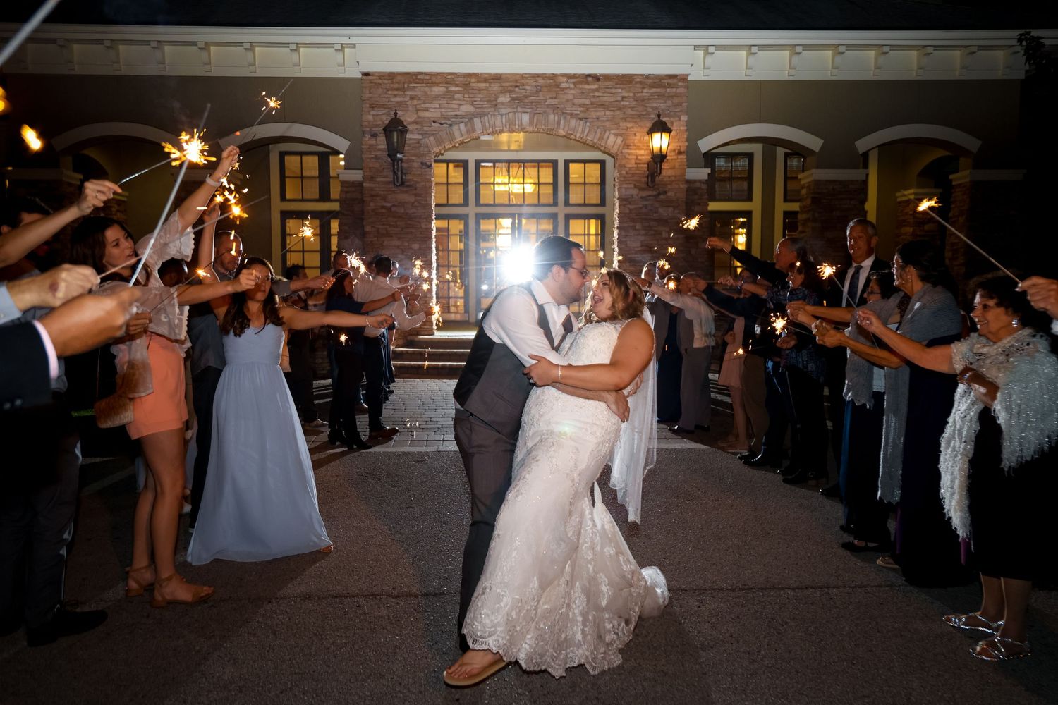 Newlyweds share their first dance under sparklers held by wedding guests outside a warmly lit brick venue at night.