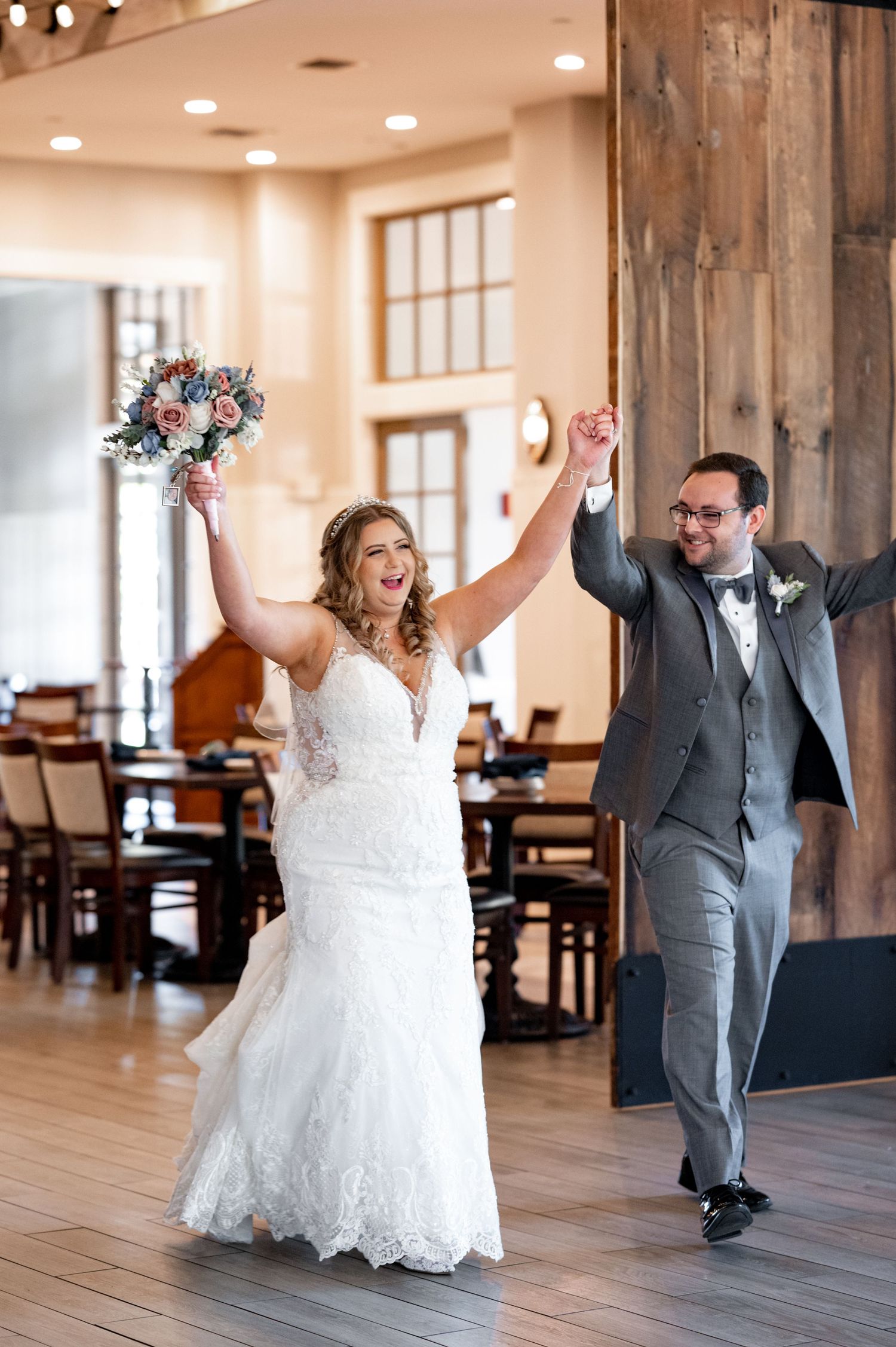 A bride in a lace gown and groom in grey suit celebrate joyfully with raised arms at a rustic wedding venue.
