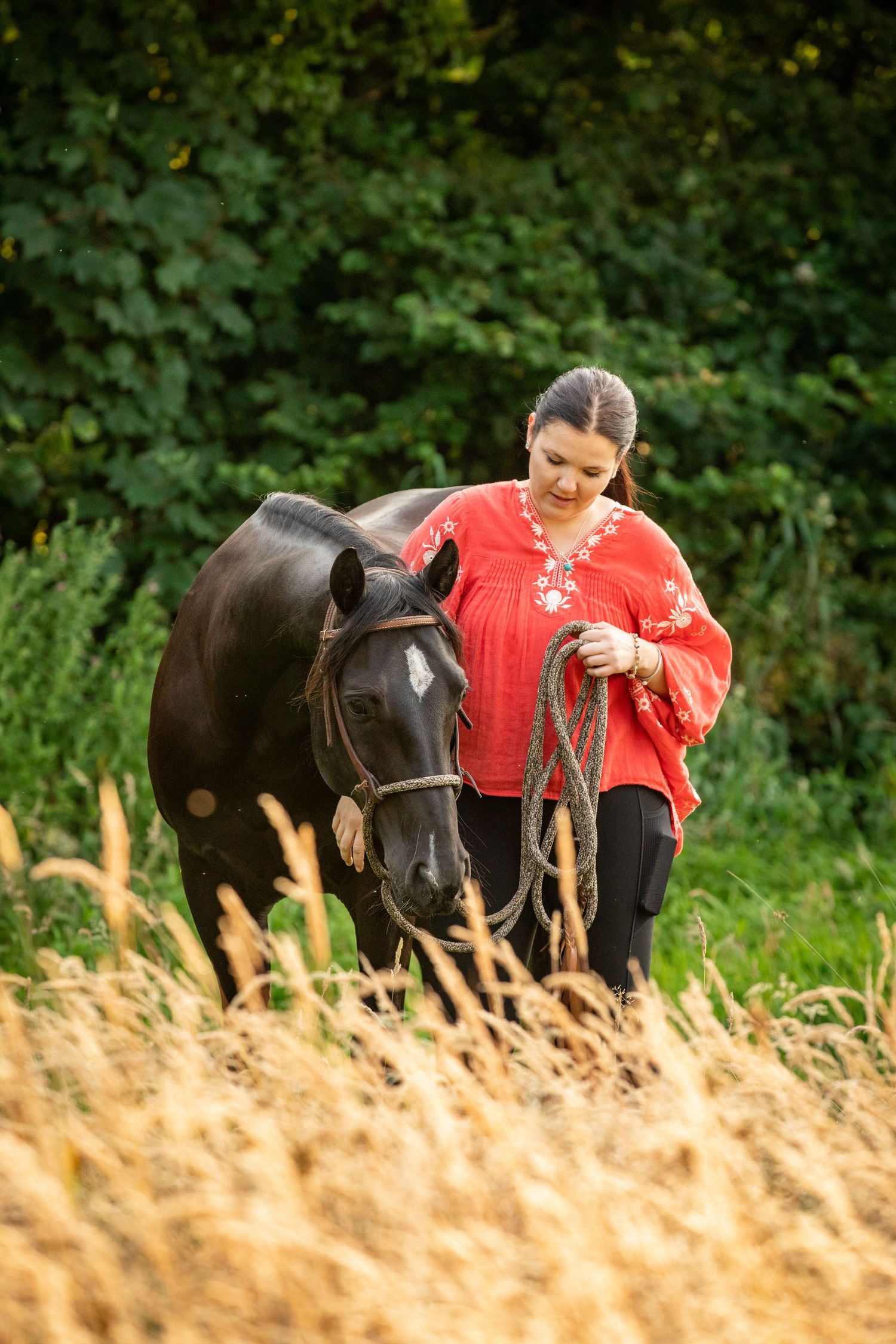 A horse and its caretaker stand together in a field with tall grass and green foliage.