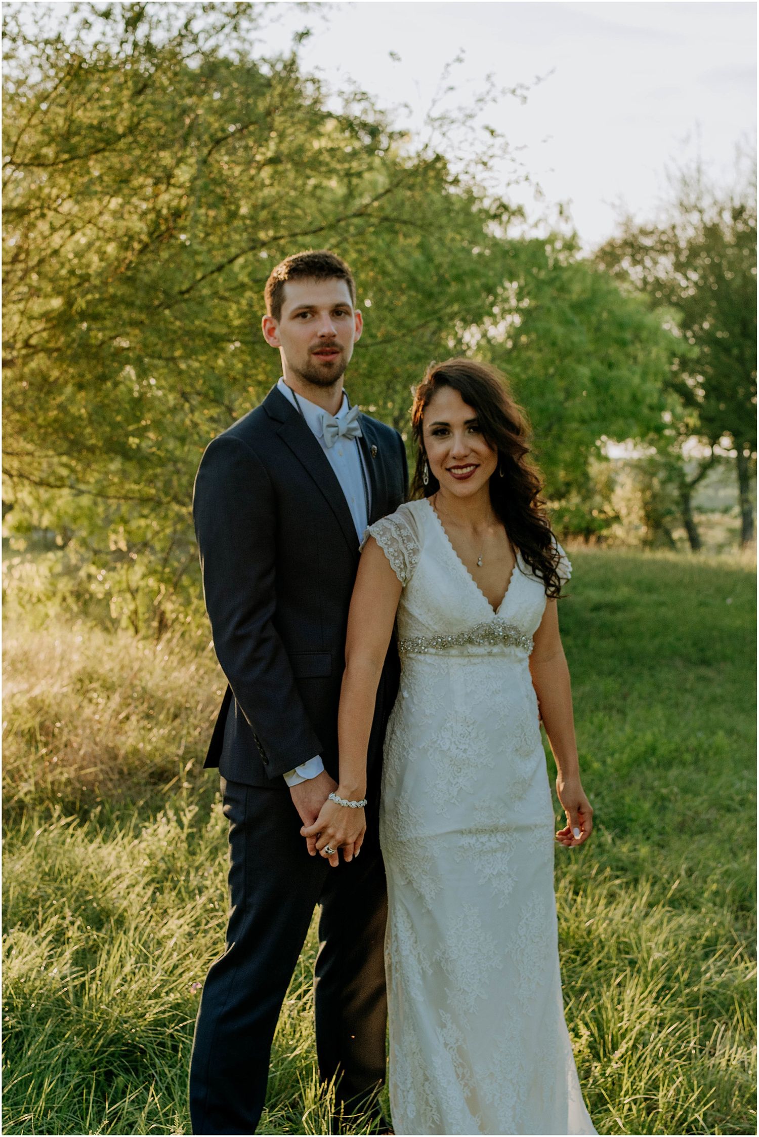 A couple poses together in elegant wedding attire against a natural outdoor backdrop.